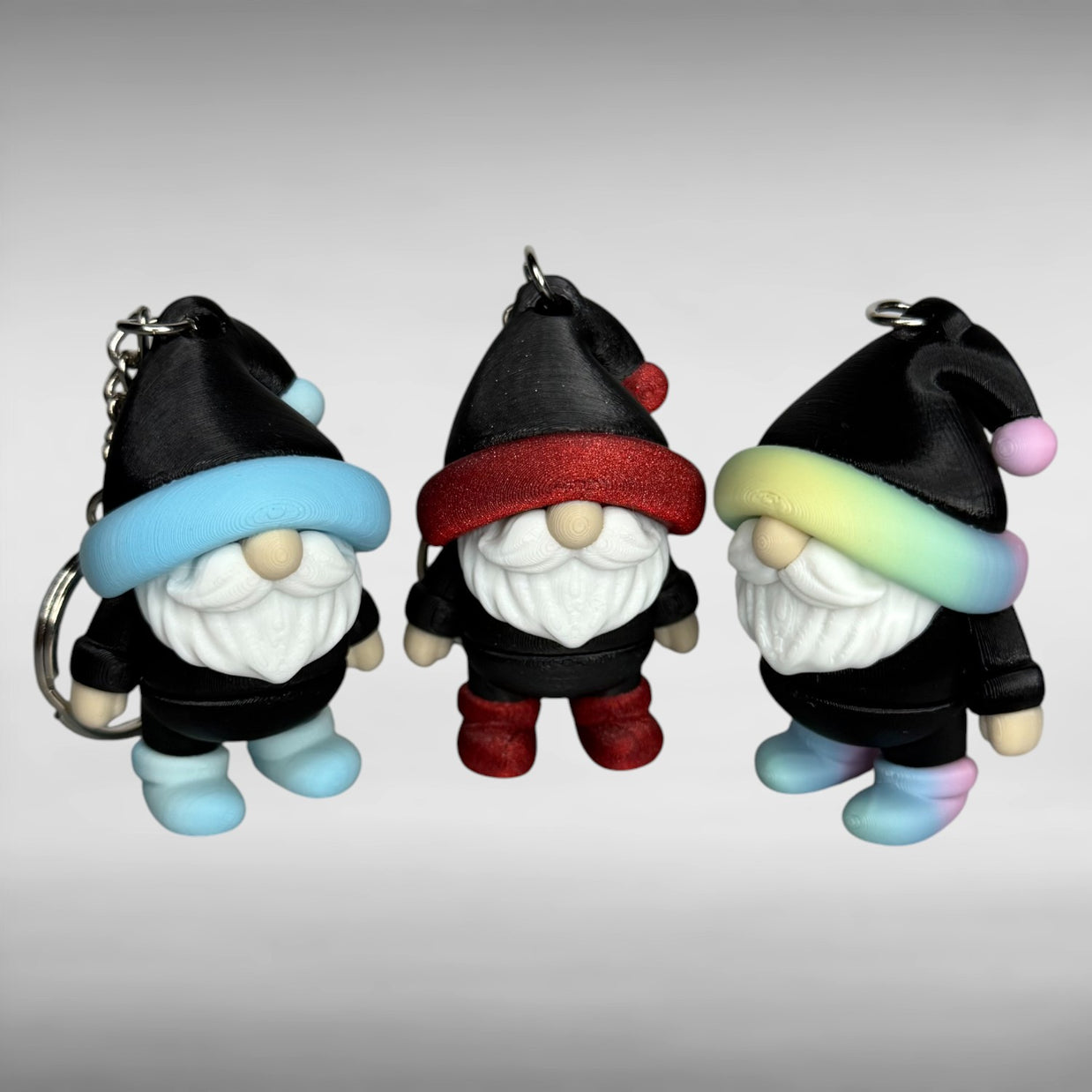 Three gnome-shaped keychains in black with different colored hats (blue/white gradient and black, shimmer red and black, and pastel rainbow gradient and black) with shoes the same color as the hats on a gray background