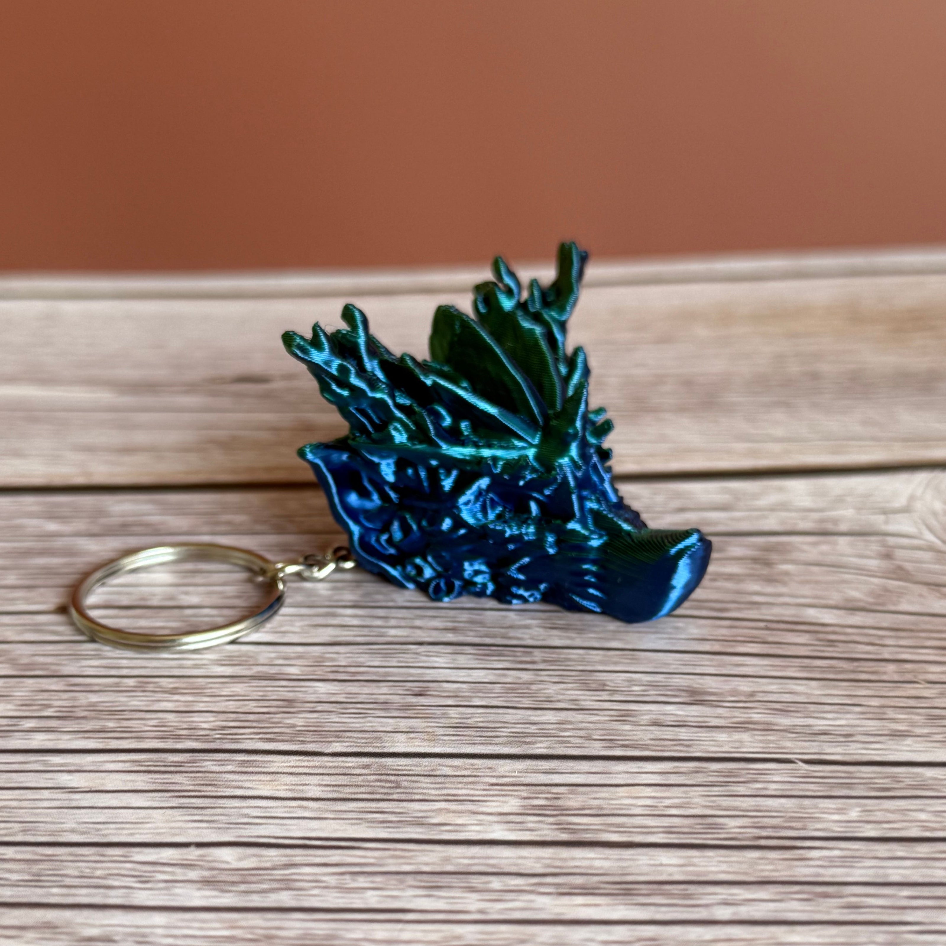 A 3D printed keychain shaped like a dragon head with coral-like details, primarily in green and blue colors, placed on a wooden surface