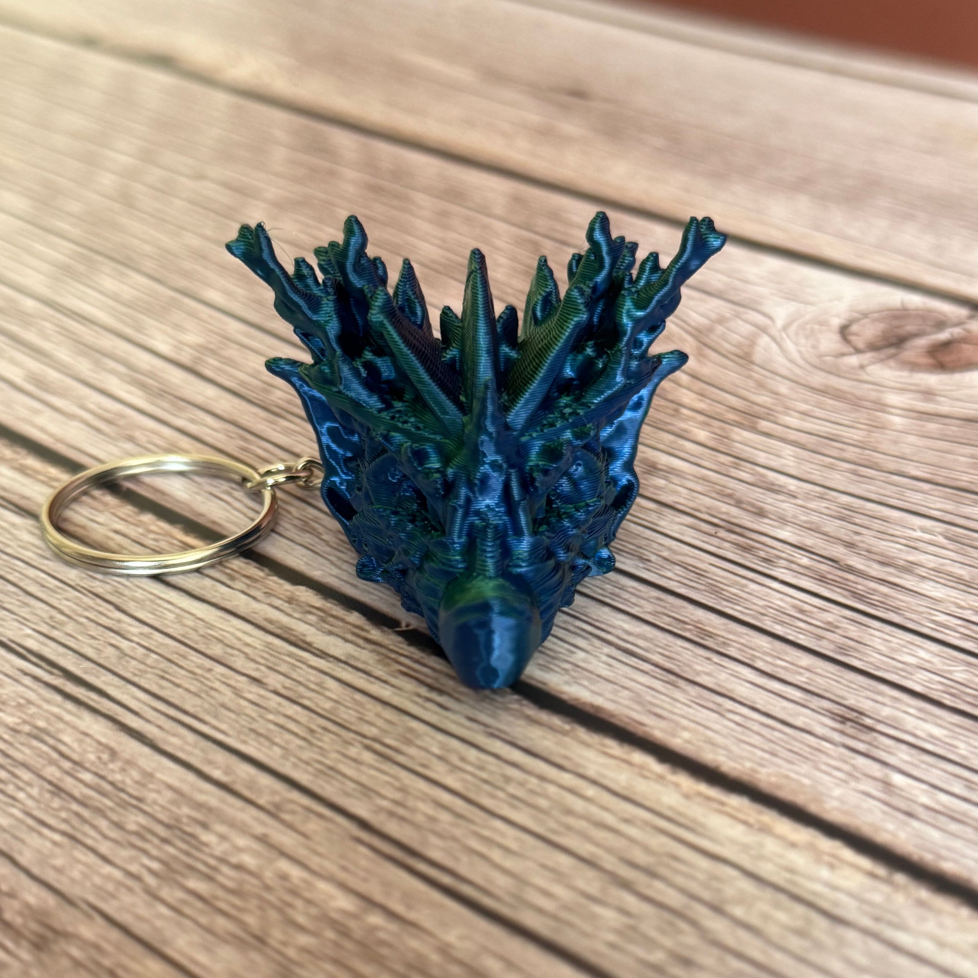 A 3D printed keychain shaped like a dragon head with coral-like details, primarily in green and blue colors, placed on a wooden surface
