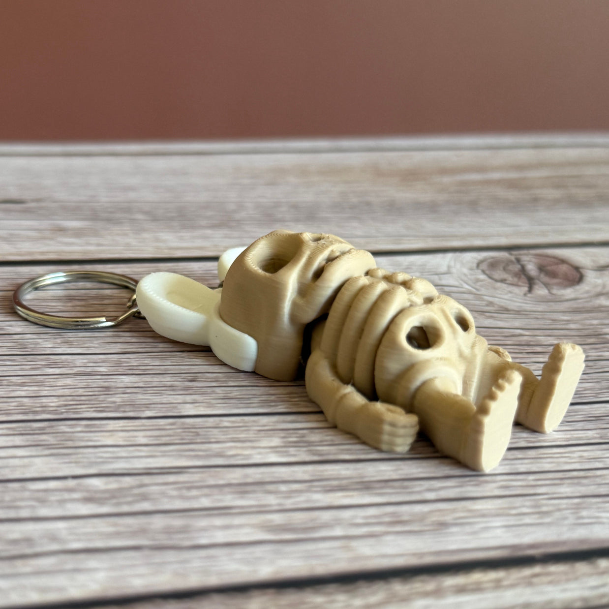 Side of a 3D printed keychain of a beige skeleton with white bunny ears on a wooden surface