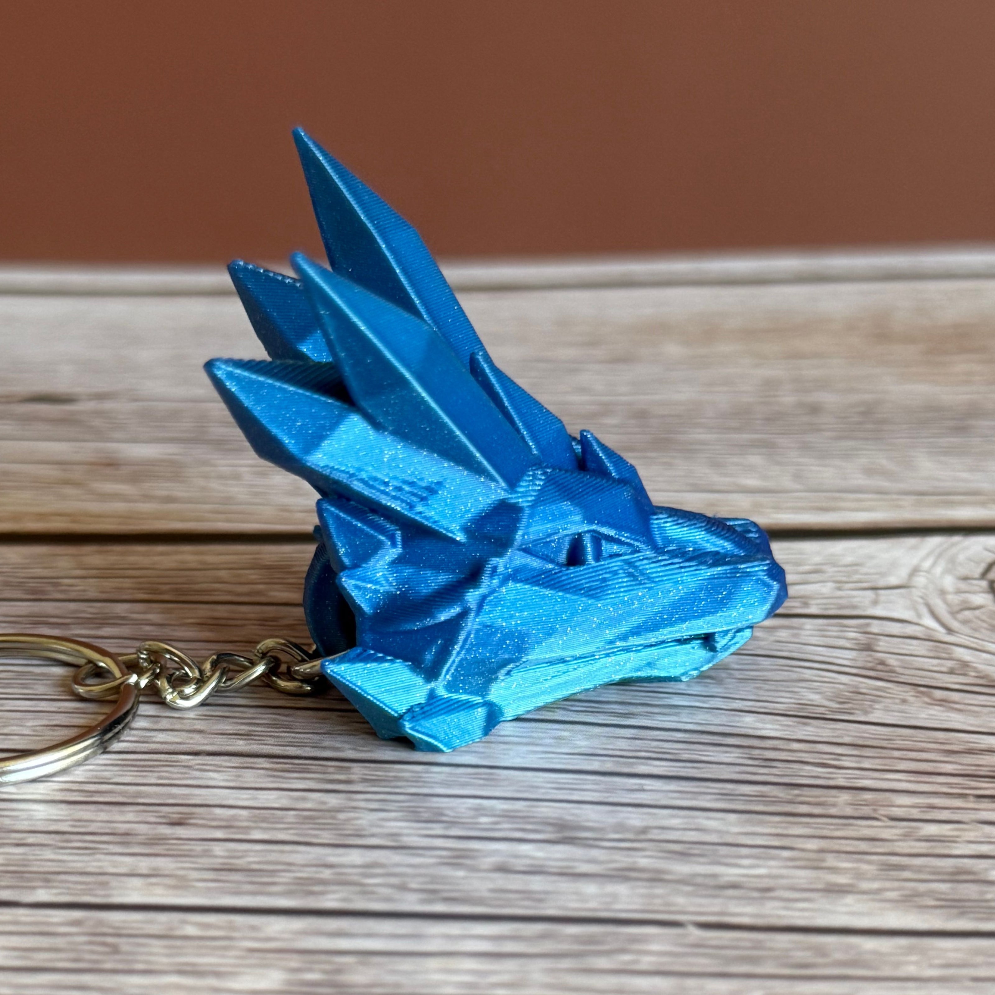 Side of a blue 3D printed keychain shaped like a dragon head with crystal-like details, placed on a wooden surface