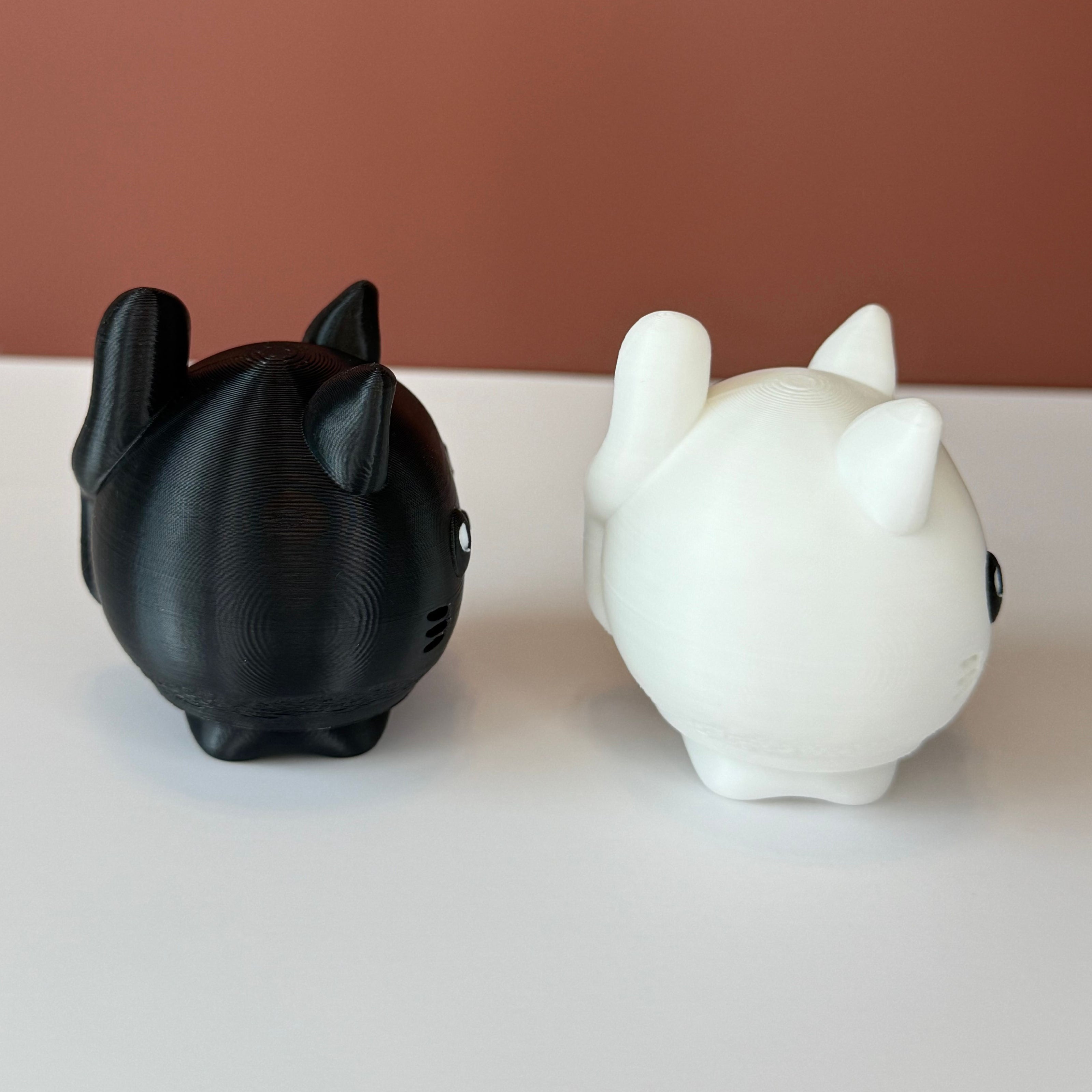 Side view of 2 cat-shaped 3D printed figures, one black and one white, on a white surface and brown backdrop