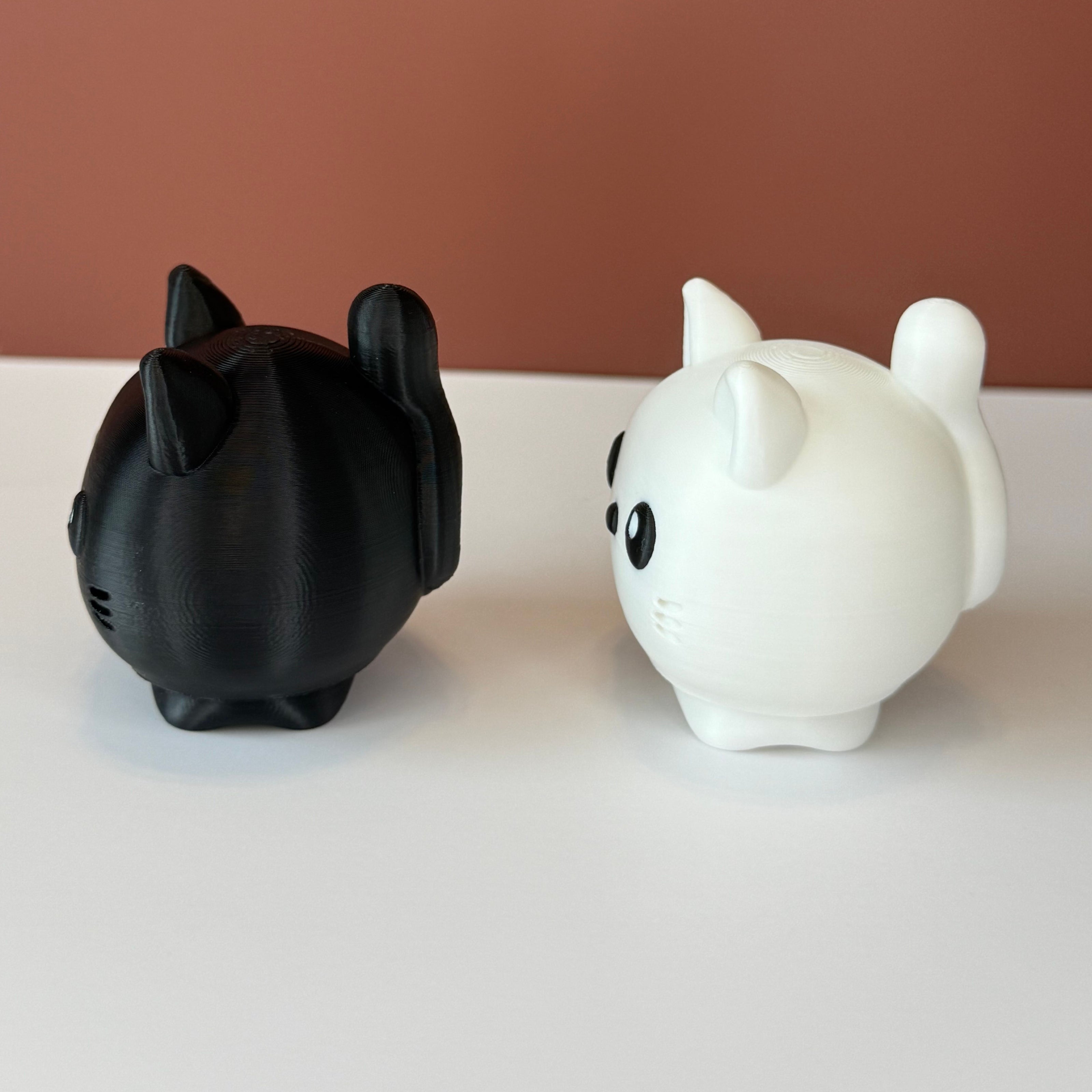 Side view of 2 cat-shaped 3D printed figures, one black and one white, on a white surface and brown backdrop