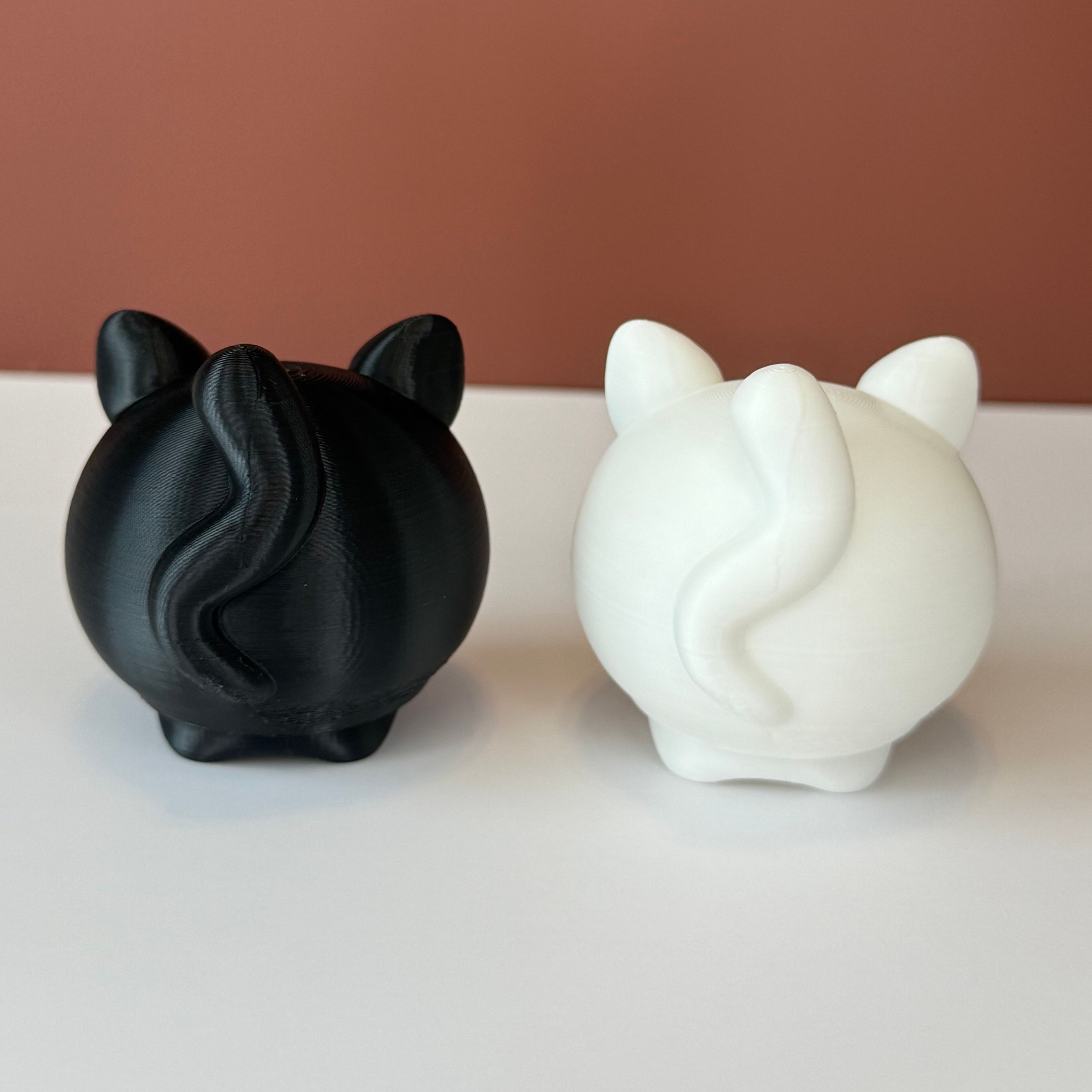 backside of 2 cat-shaped 3D printed figures, one black and one white, on a white surface and brown backdrop