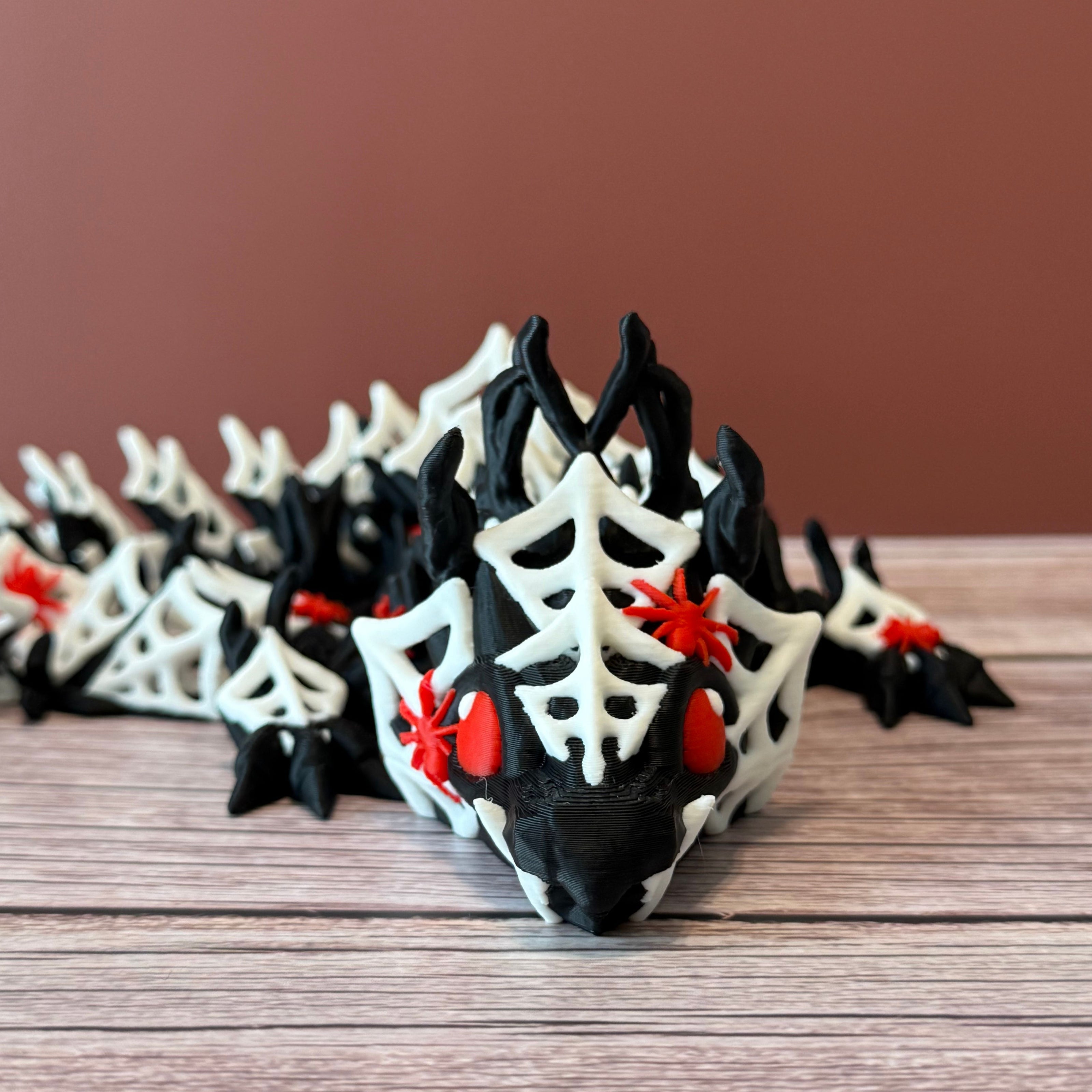 3D printed black dragon with white spider webs and red spiders across the body, placed on a wooden surface with a brown backdrop