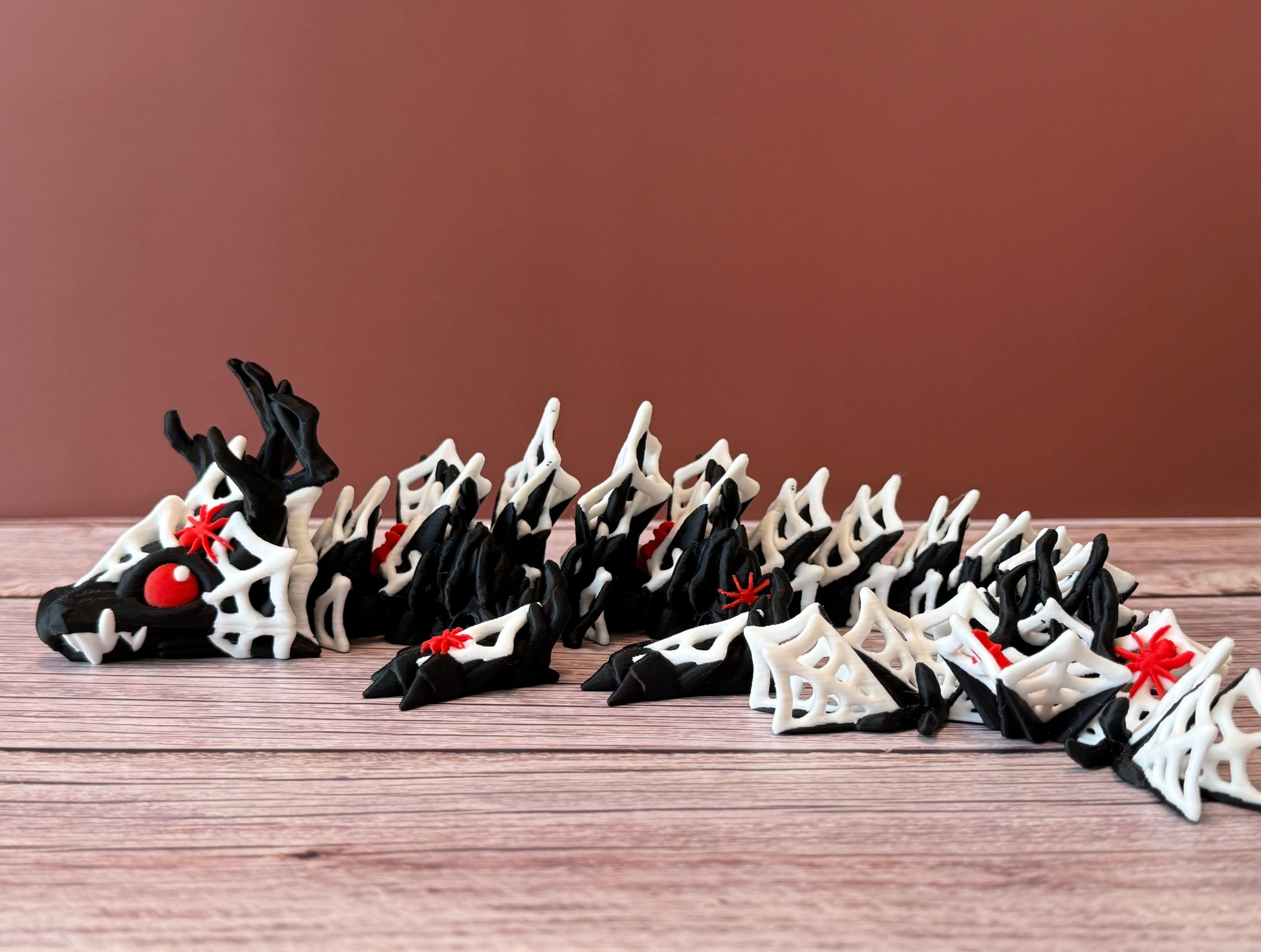 3D printed black dragon with white spider webs and red spiders across the body, placed on a wooden surface with a brown backdrop