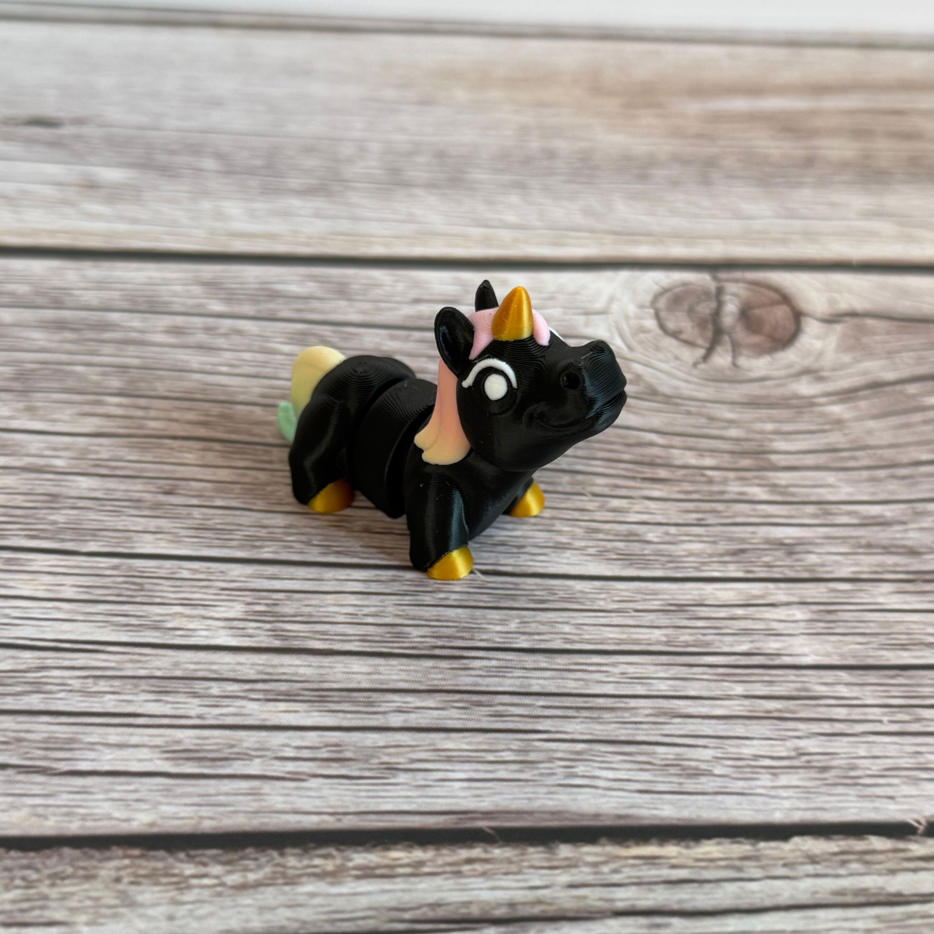 A small 3D printed articulated mini unicorn toy with articulated segments, black body, rainbow hair, and golden hooves and horn on a wooden surface