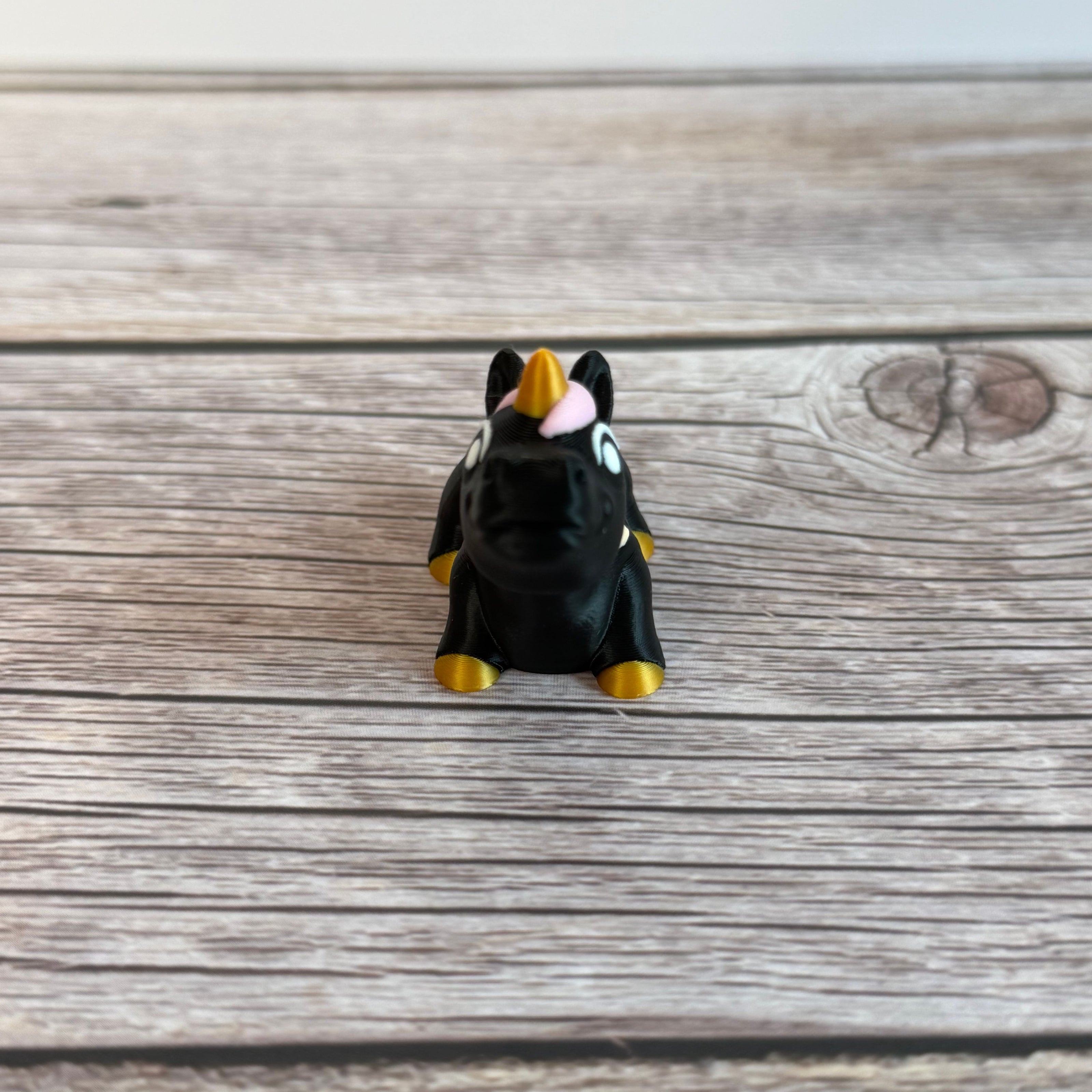 A small 3D printed articulated mini unicorn toy with articulated segments, black body, rainbow hair, and golden hooves and horn on a wooden surface
