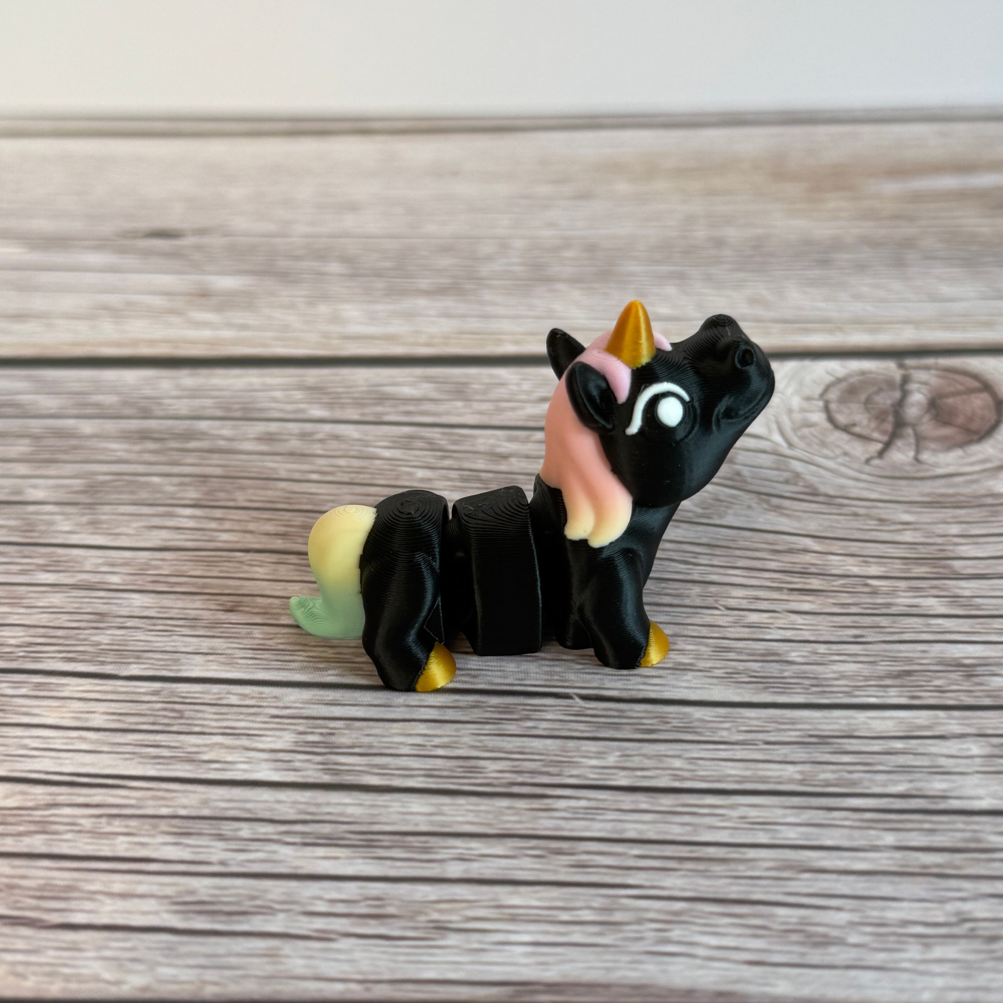 A small 3D printed articulated mini unicorn toy with articulated segments, black body, rainbow hair, and golden hooves and horn on a wooden surface