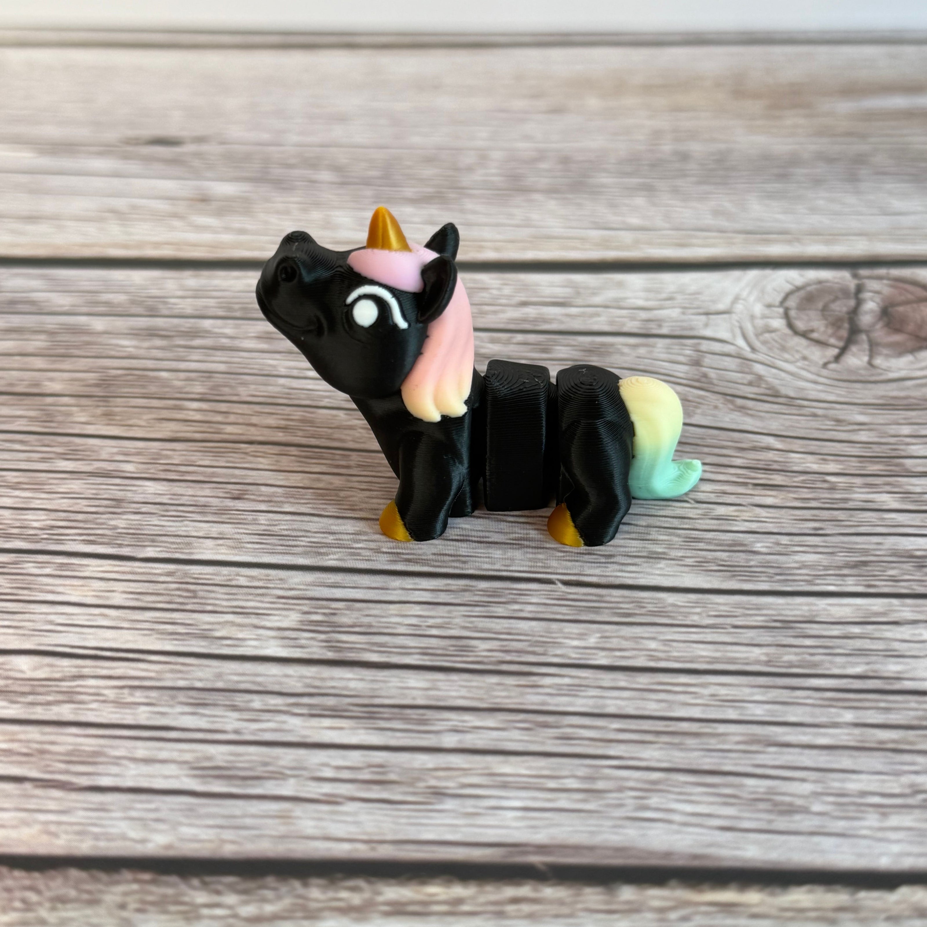 A small 3D printed articulated mini unicorn toy with articulated segments, black body, rainbow hair, and golden hooves and horn on a wooden surface