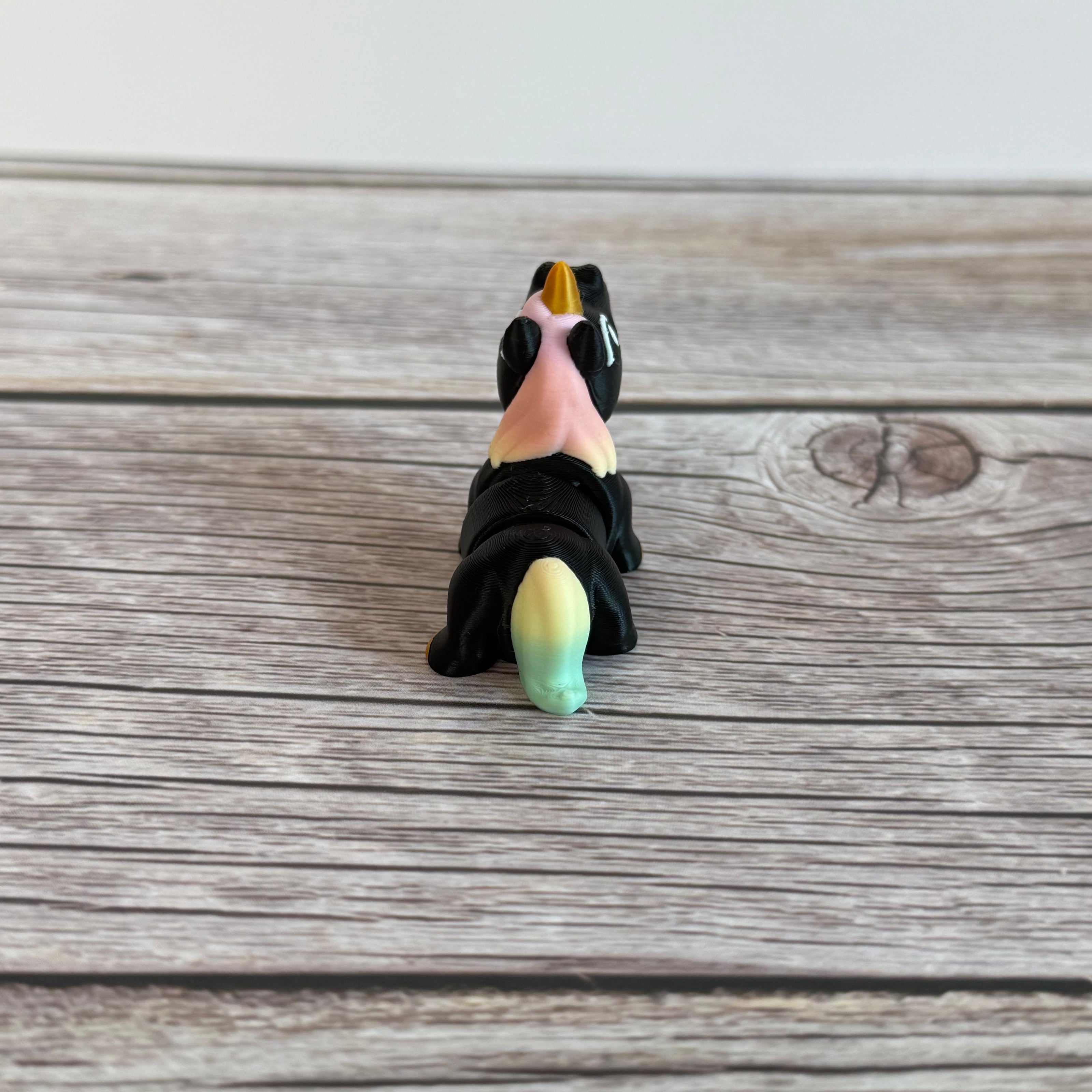 A small 3D printed articulated mini unicorn toy with articulated segments, black body, rainbow hair, and golden hooves and horn on a wooden surface