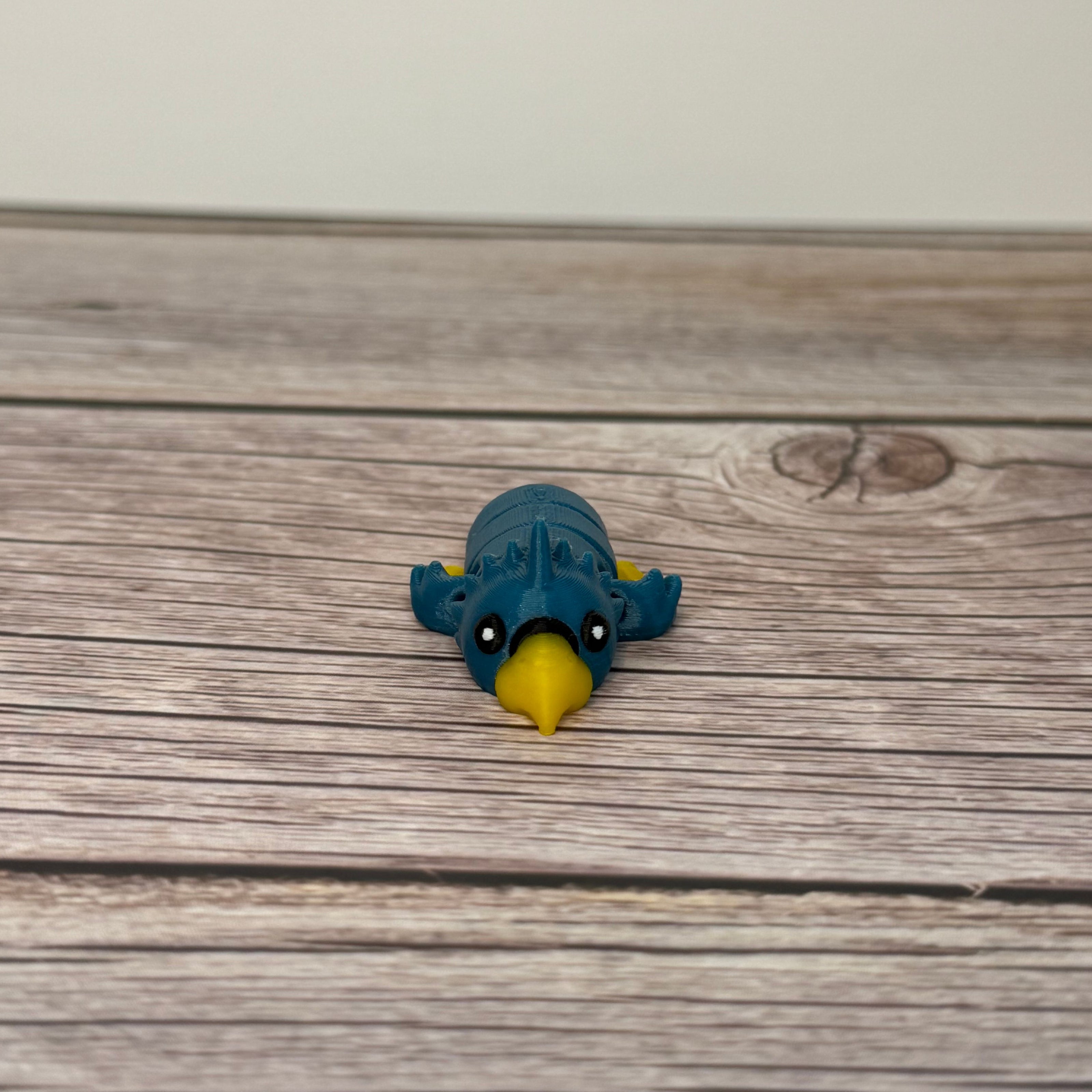 A small 3D printed blue and yellow articulated mini parrot toy placed on a wooden surface.