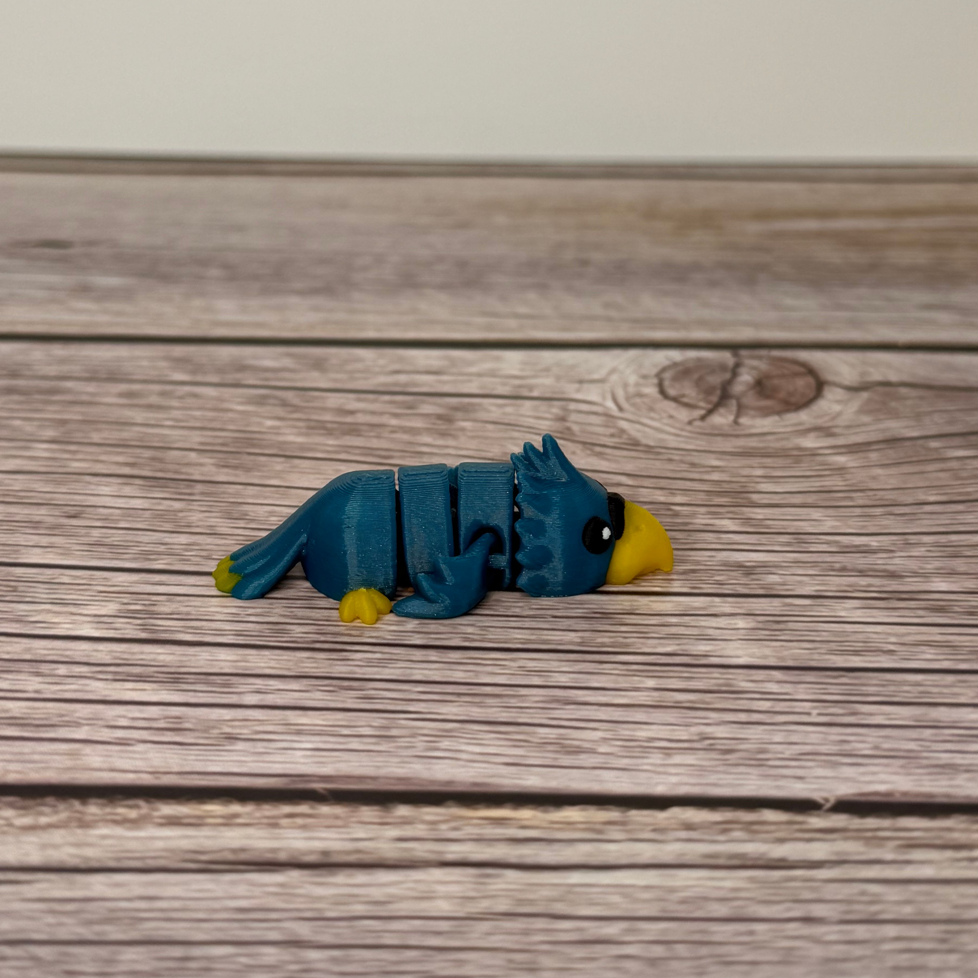 A small 3D printed blue and yellow articulated mini parrot toy placed on a wooden surface.