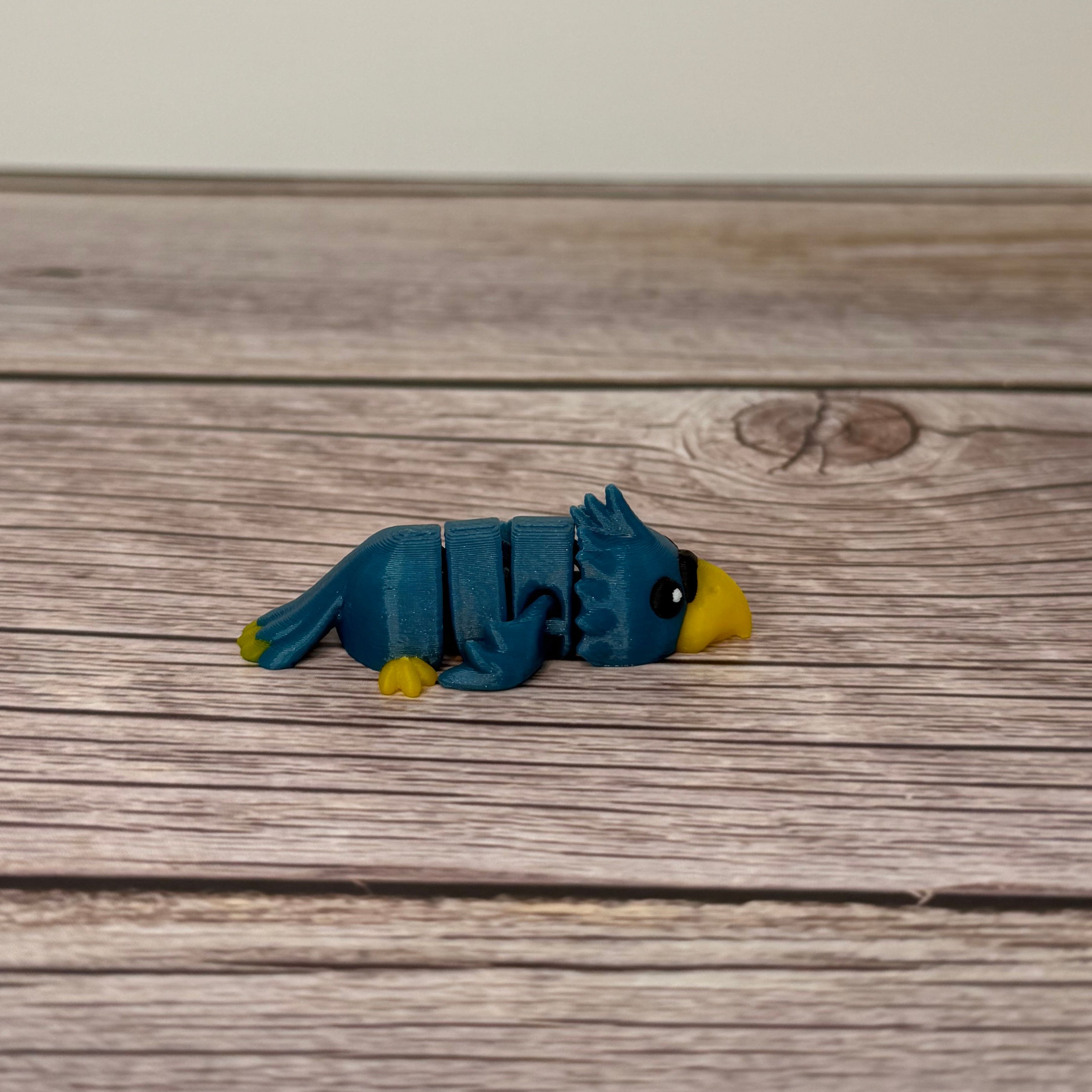A small 3D printed blue and yellow articulated mini parrot toy placed on a wooden surface.