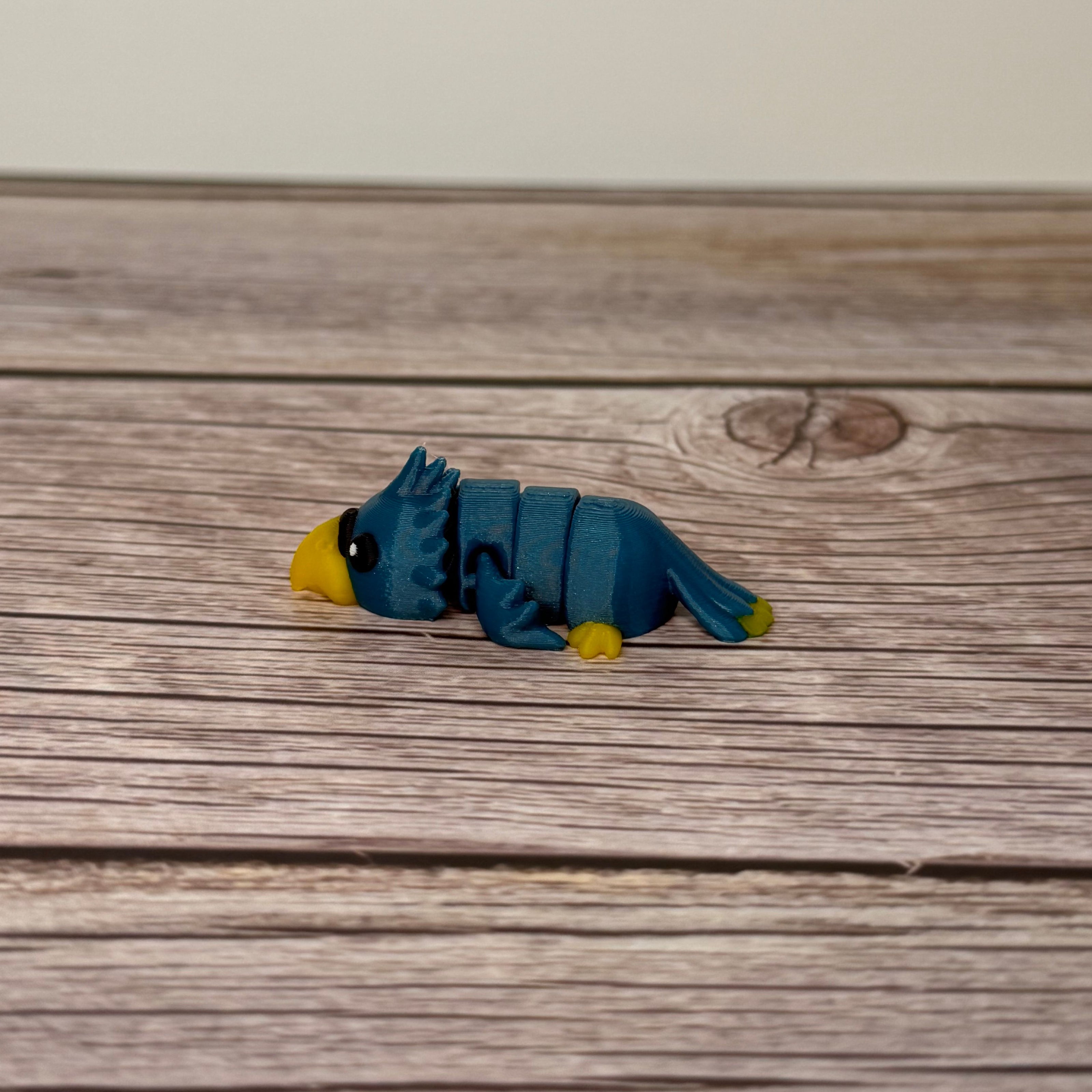 A small 3D printed blue and yellow articulated mini parrot toy placed on a wooden surface.