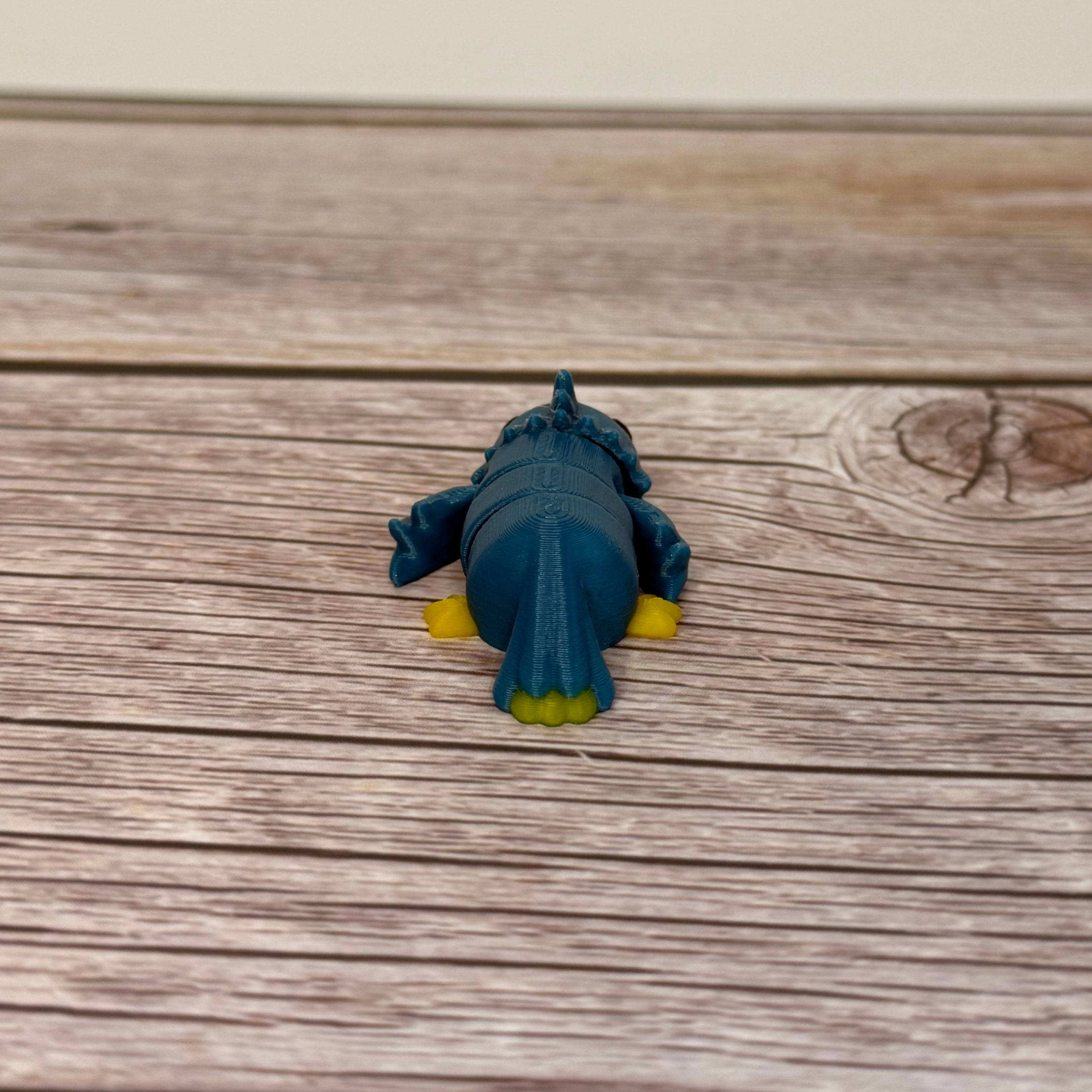 A small 3D printed blue and yellow articulated mini parrot toy placed on a wooden surface.