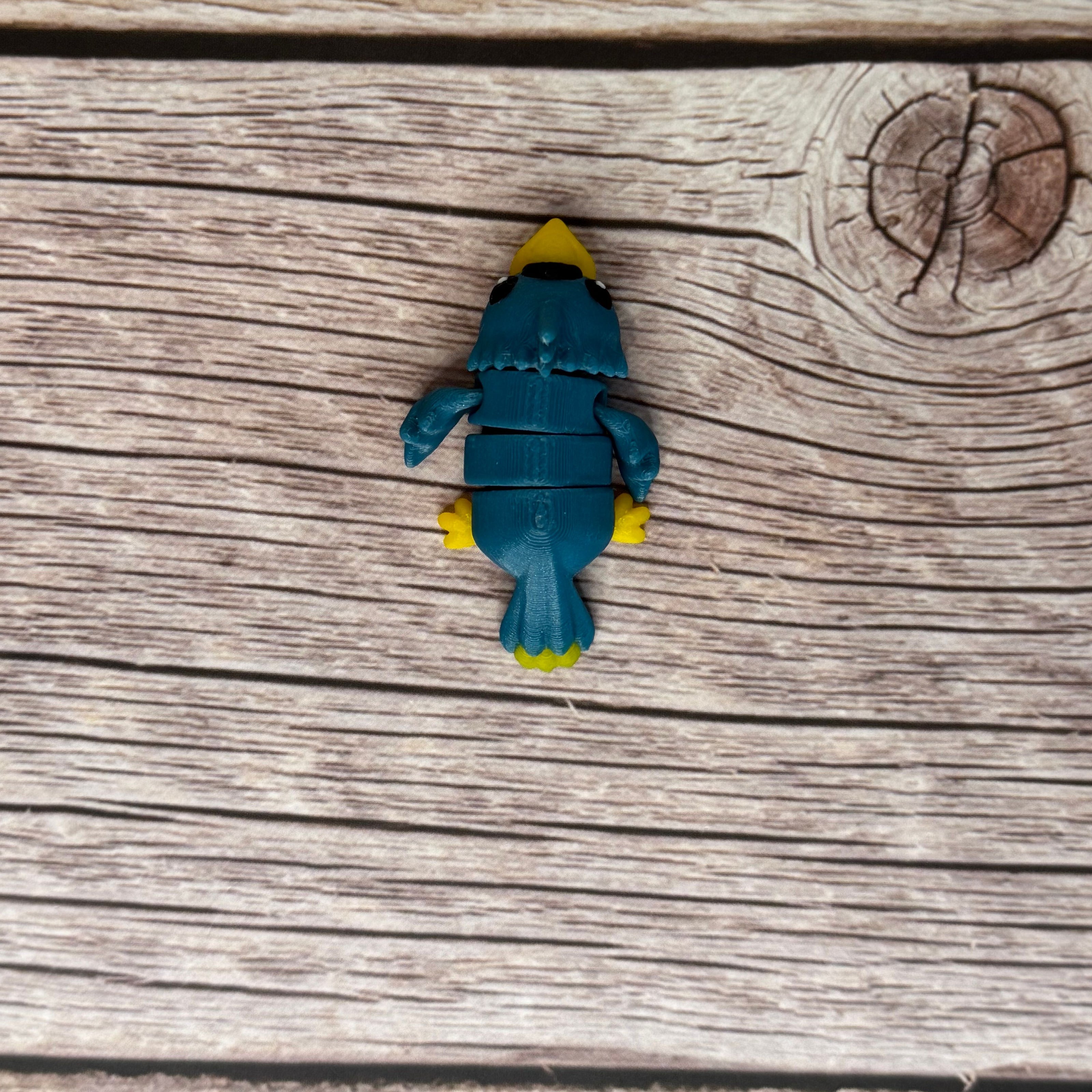 A small 3D printed blue and yellow articulated mini parrot toy placed on a wooden surface.