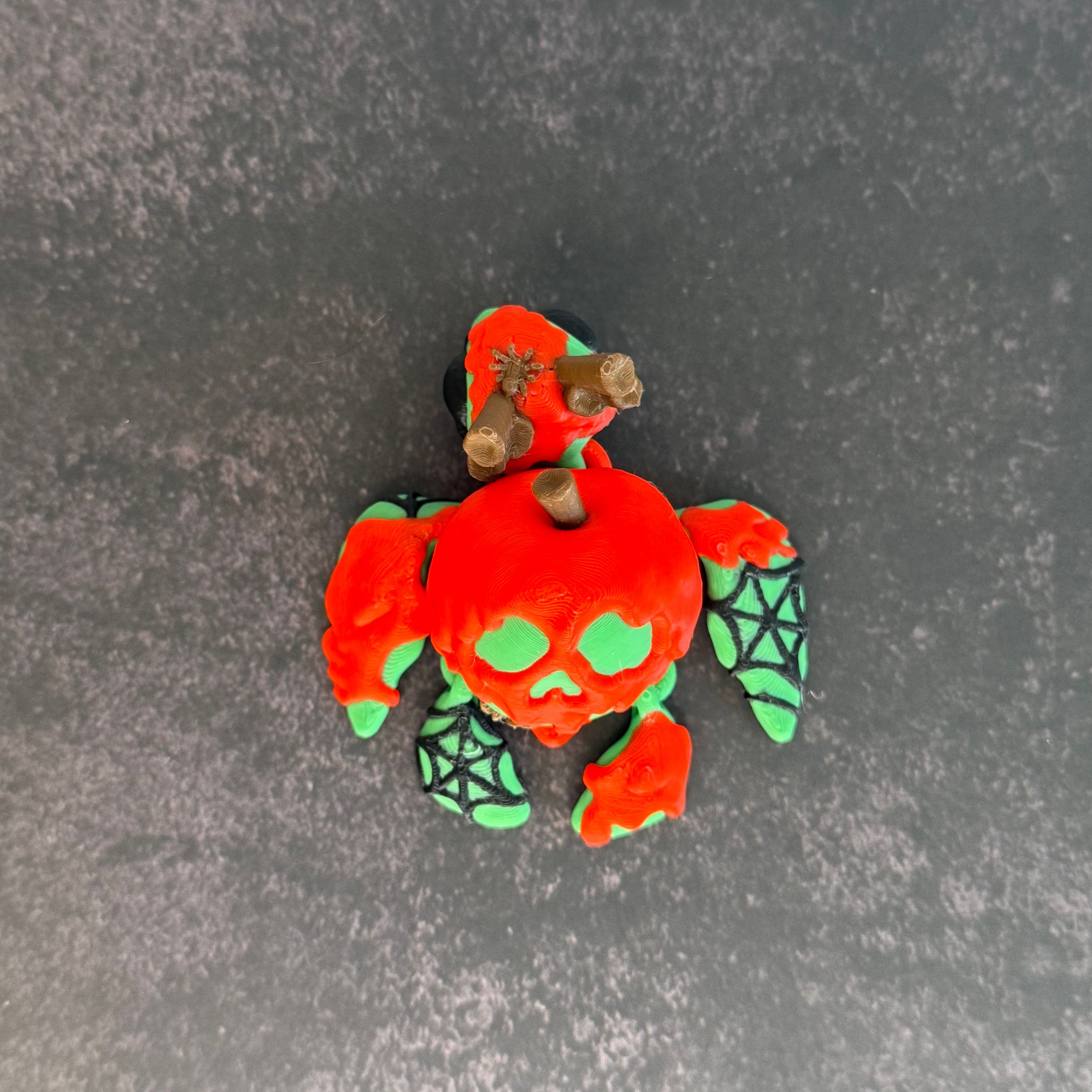 3D printed articulated turtle toy with a toxic apple-inspired shell in red and green, with a black background.