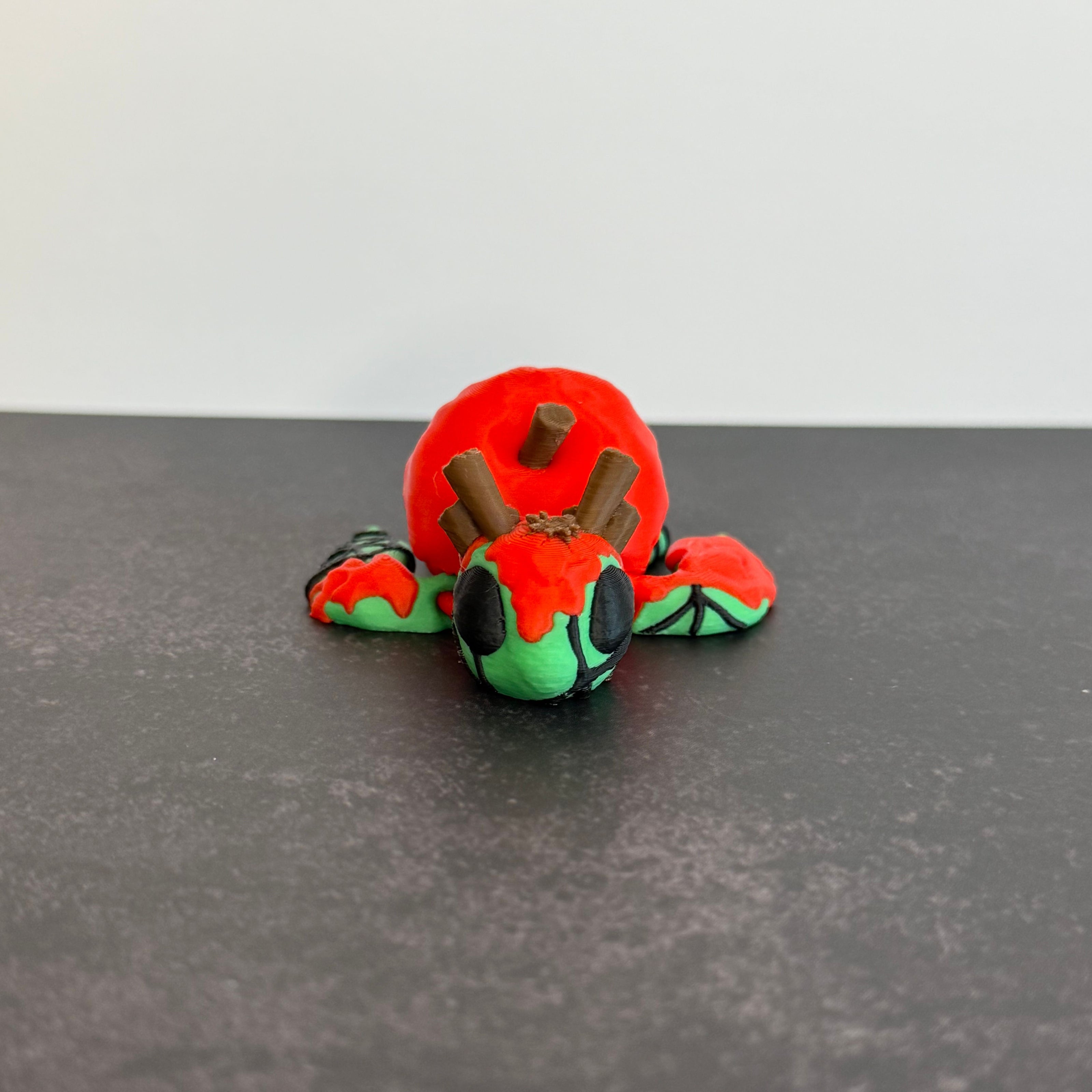 3D printed articulated turtle toy with a toxic apple-inspired shell in red and green, with a black background.