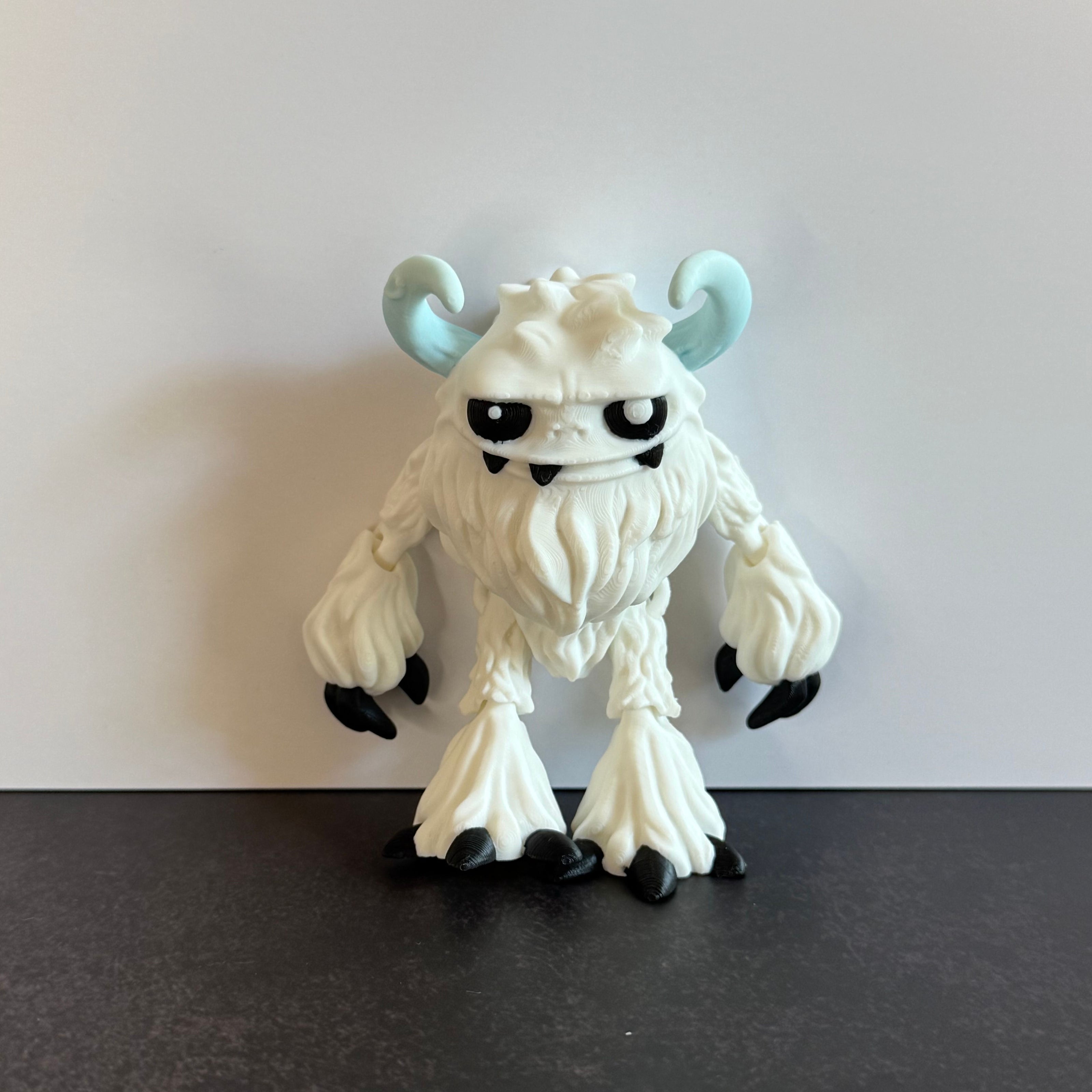 3d-printed toy figure of a yeti creature with white hairy body, black eyes, and blue horns on a black and white background