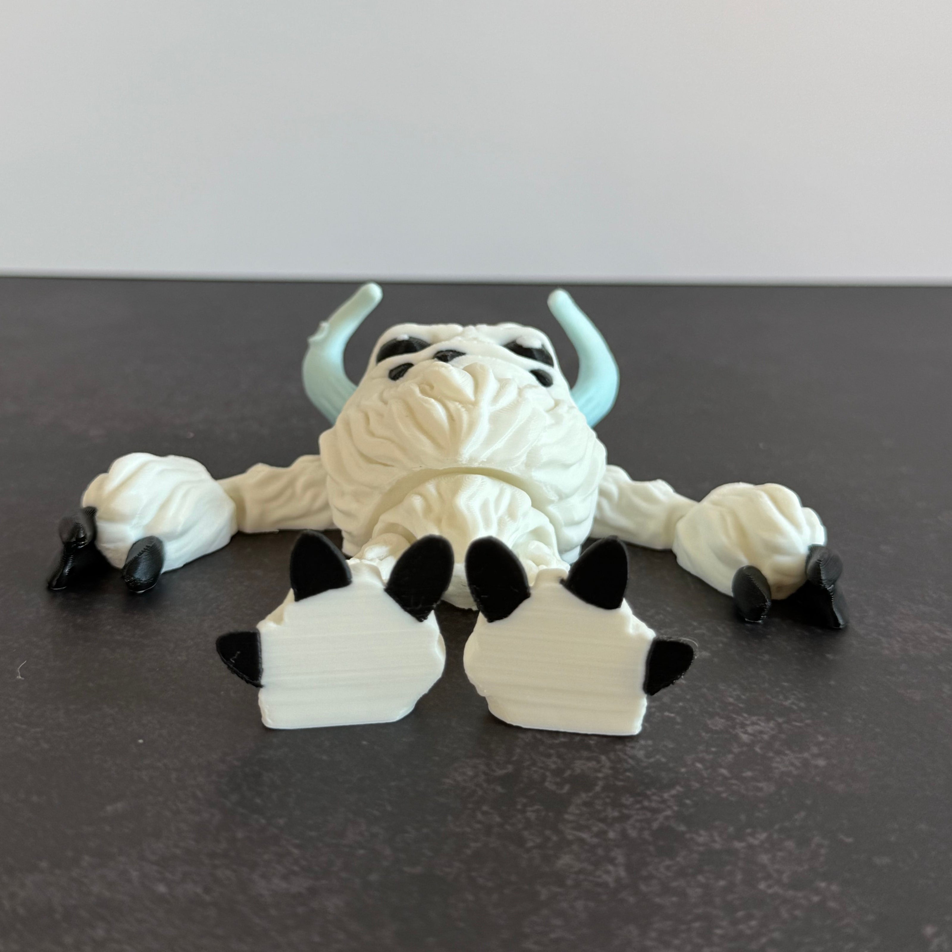 3d-printed toy figure of a yeti creature with white hairy body, black eyes, and blue horns on a black and white background