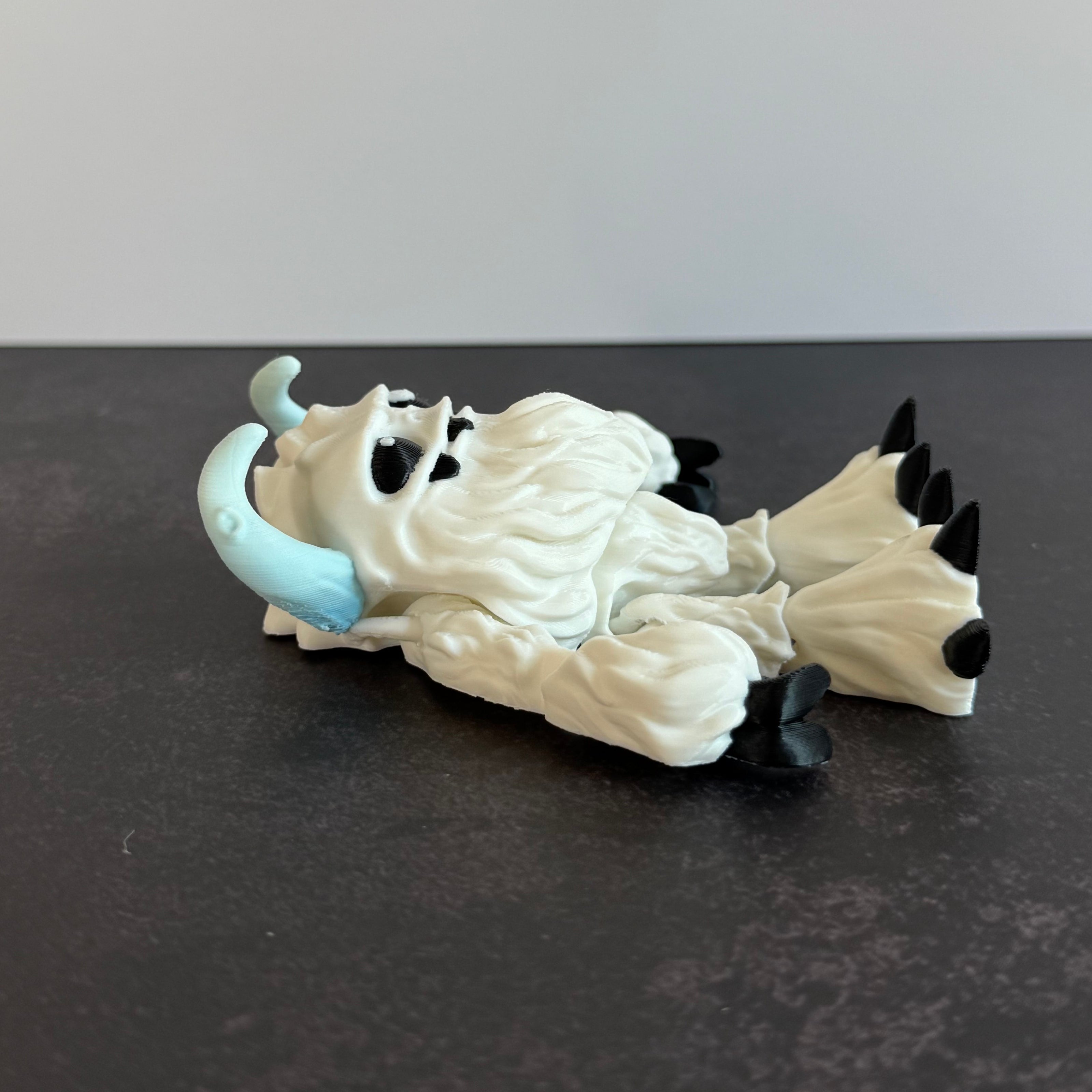3d-printed toy figure of a yeti creature with white hairy body, black eyes, and blue horns on a black and white background