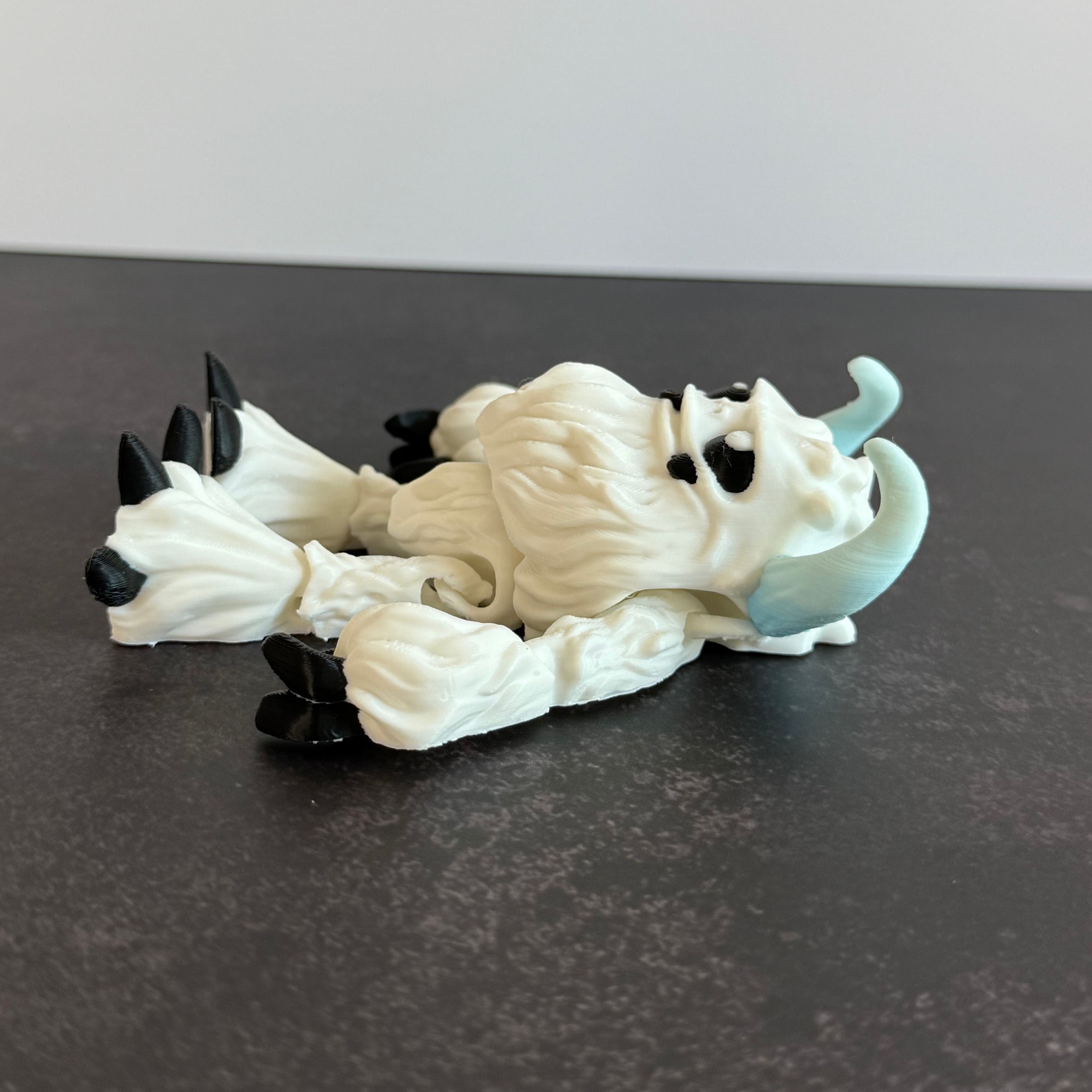 3d-printed toy figure of a yeti creature with white hairy body, black eyes, and blue horns on a black and white background