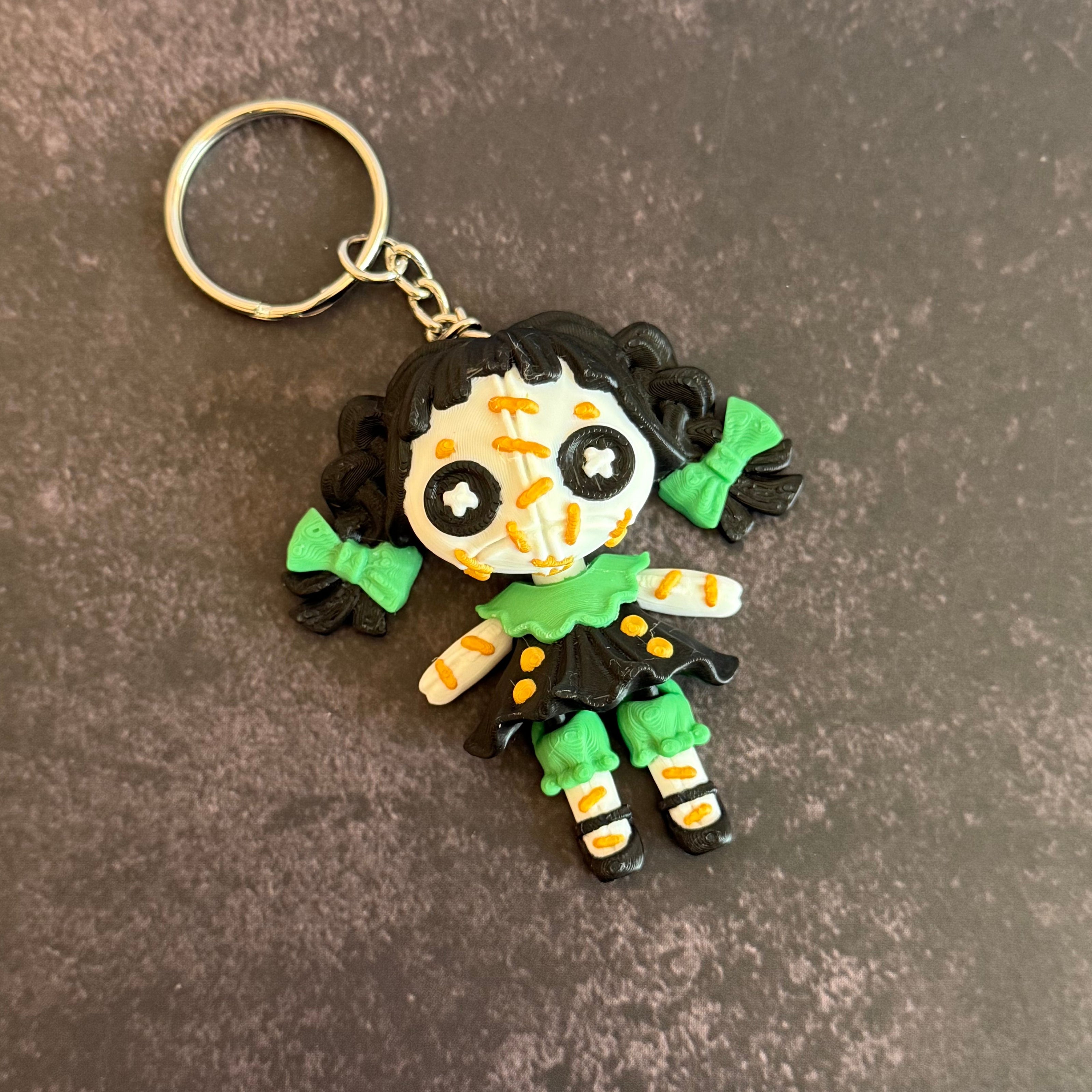 A 3D printed keychain depicting a small, creepy doll with black hair, green accents, and an orange stitch pattern. The keychain is attached to a metal ring.