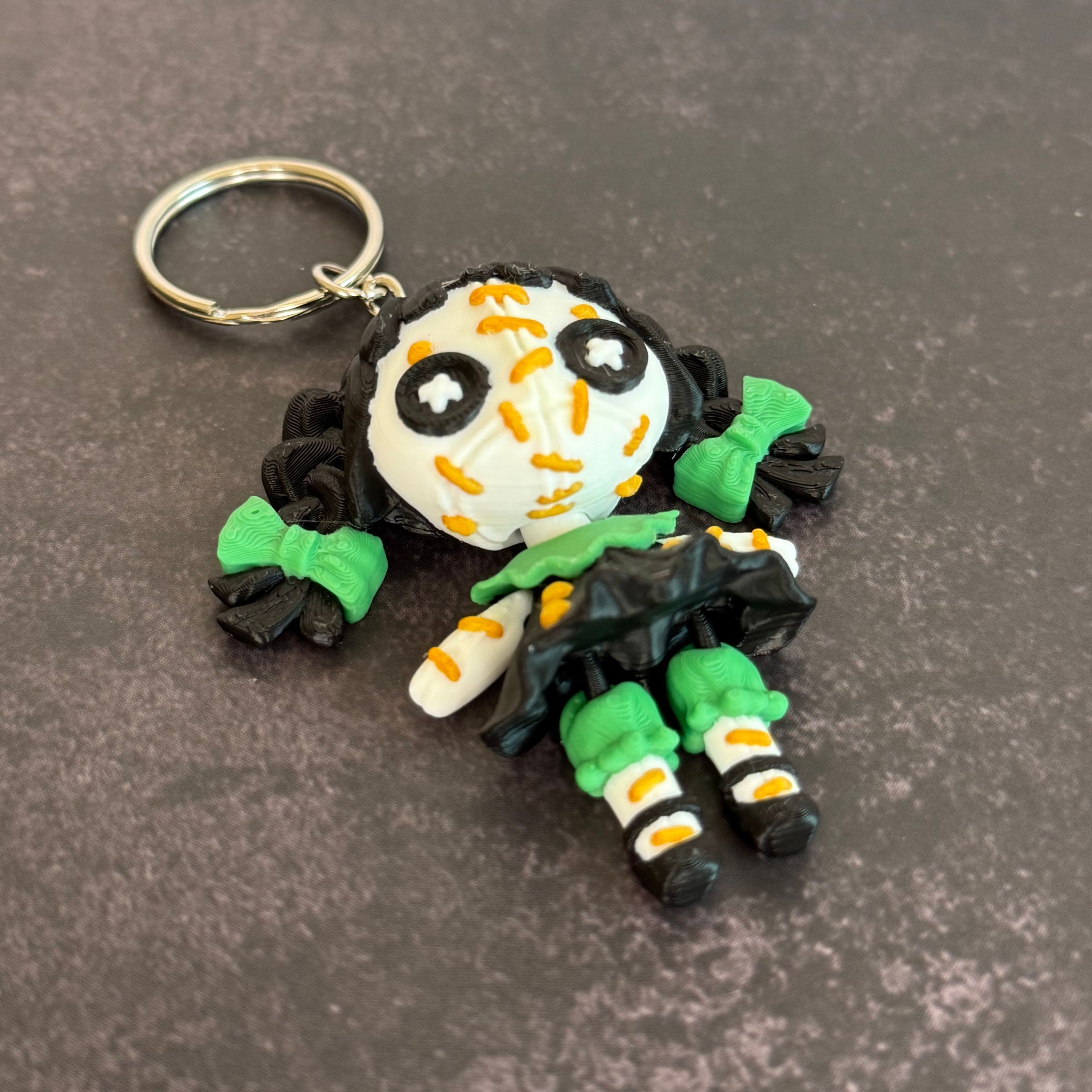 A 3D printed keychain depicting a small, creepy doll with black hair, green accents, and an orange stitch pattern. The keychain is attached to a metal ring.