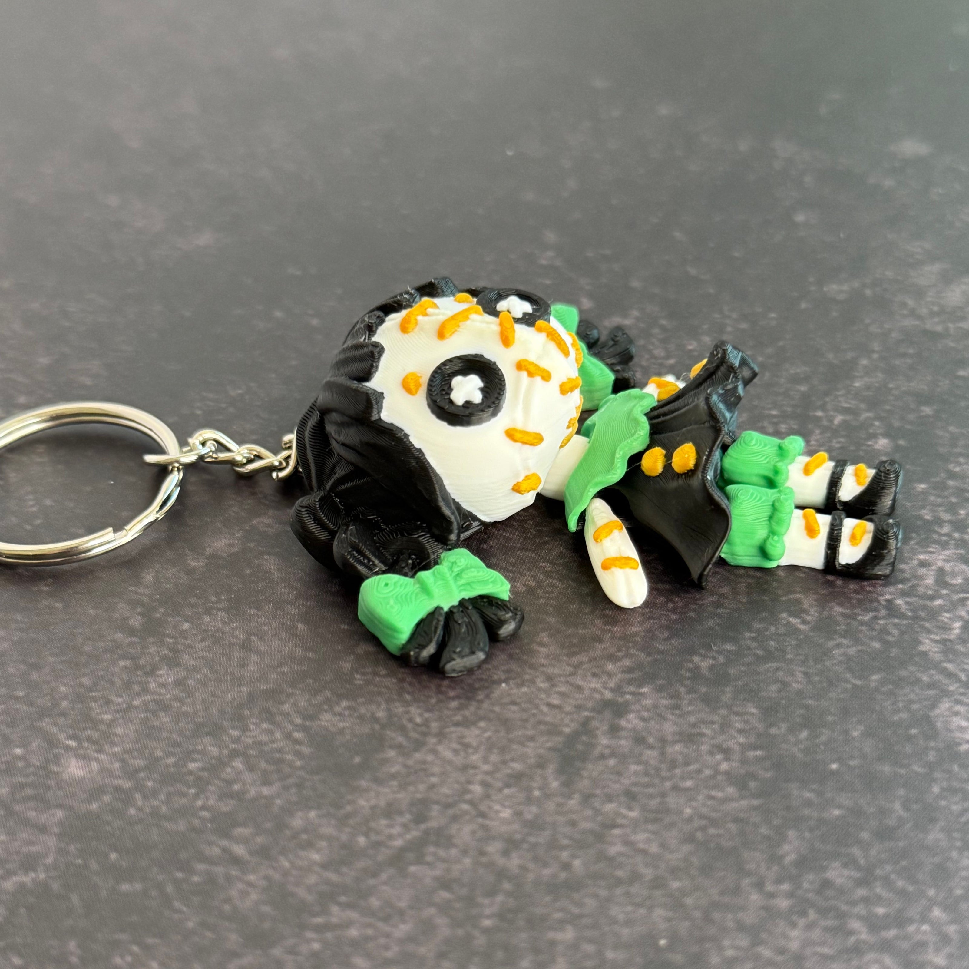 A 3D printed keychain depicting a small, creepy doll with black hair, green accents, and an orange stitch pattern. The keychain is attached to a metal ring.