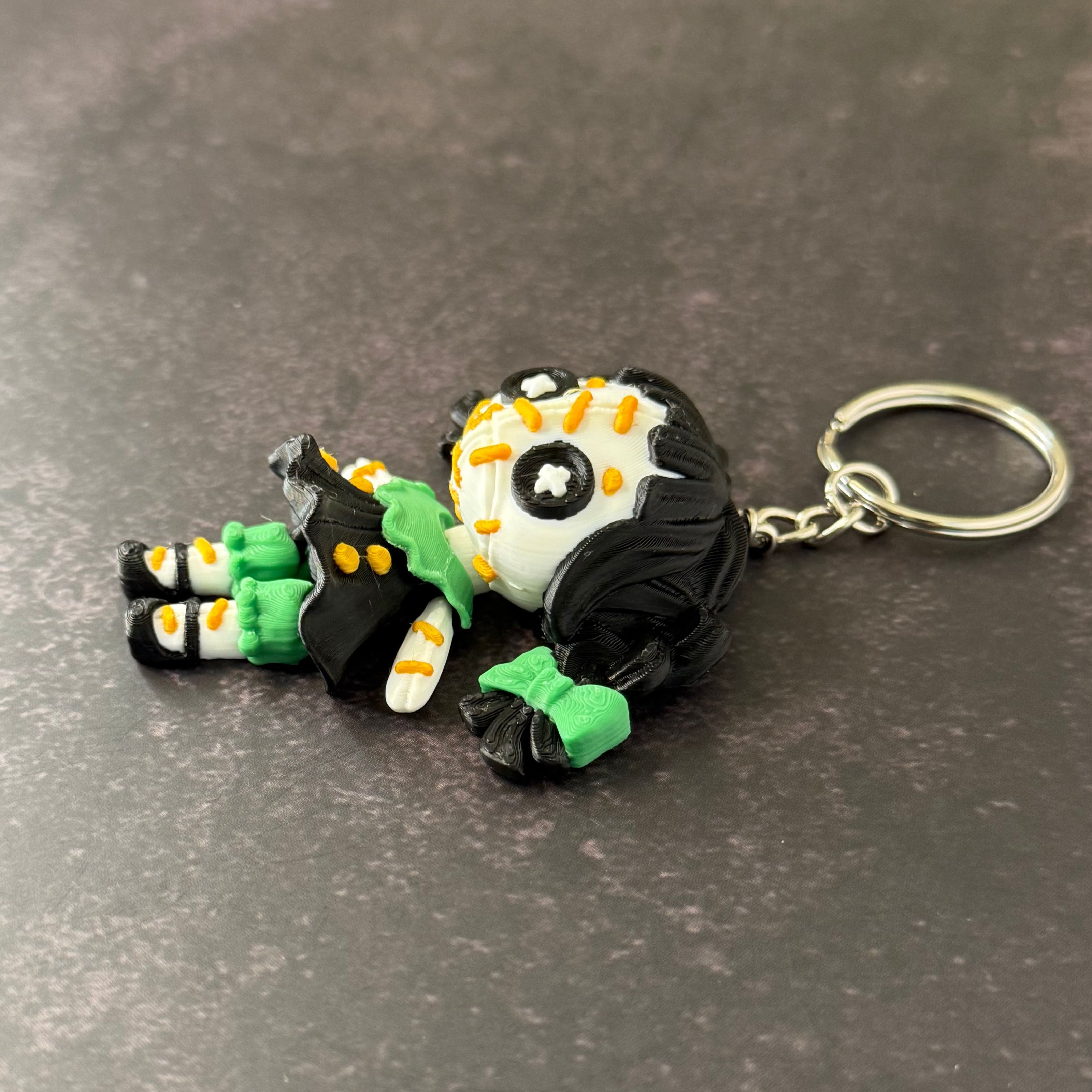 A 3D printed keychain depicting a small, creepy doll with black hair, green accents, and an orange stitch pattern. The keychain is attached to a metal ring.