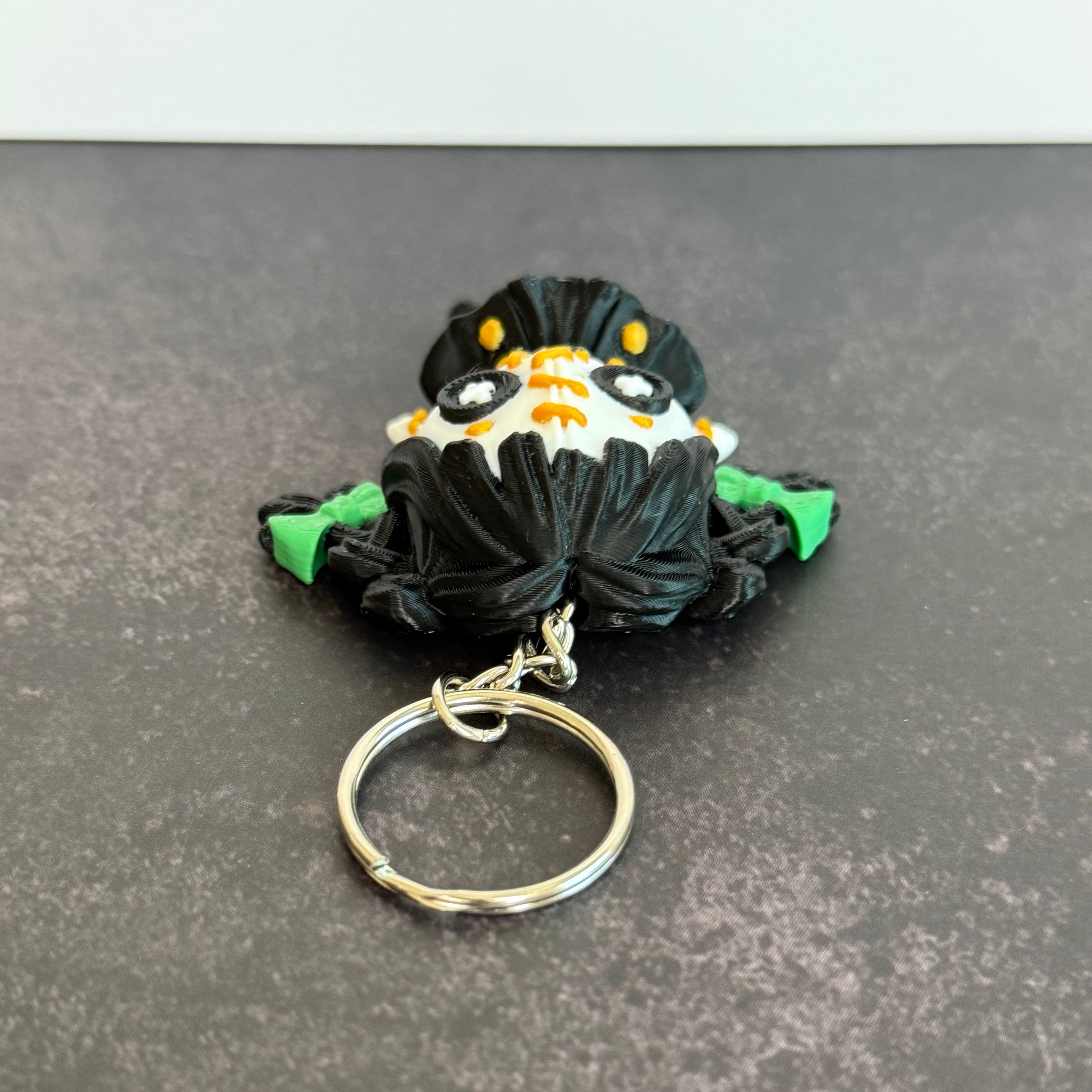 A 3D printed keychain depicting a small, creepy doll with black hair, green accents, and an orange stitch pattern. The keychain is attached to a metal ring.