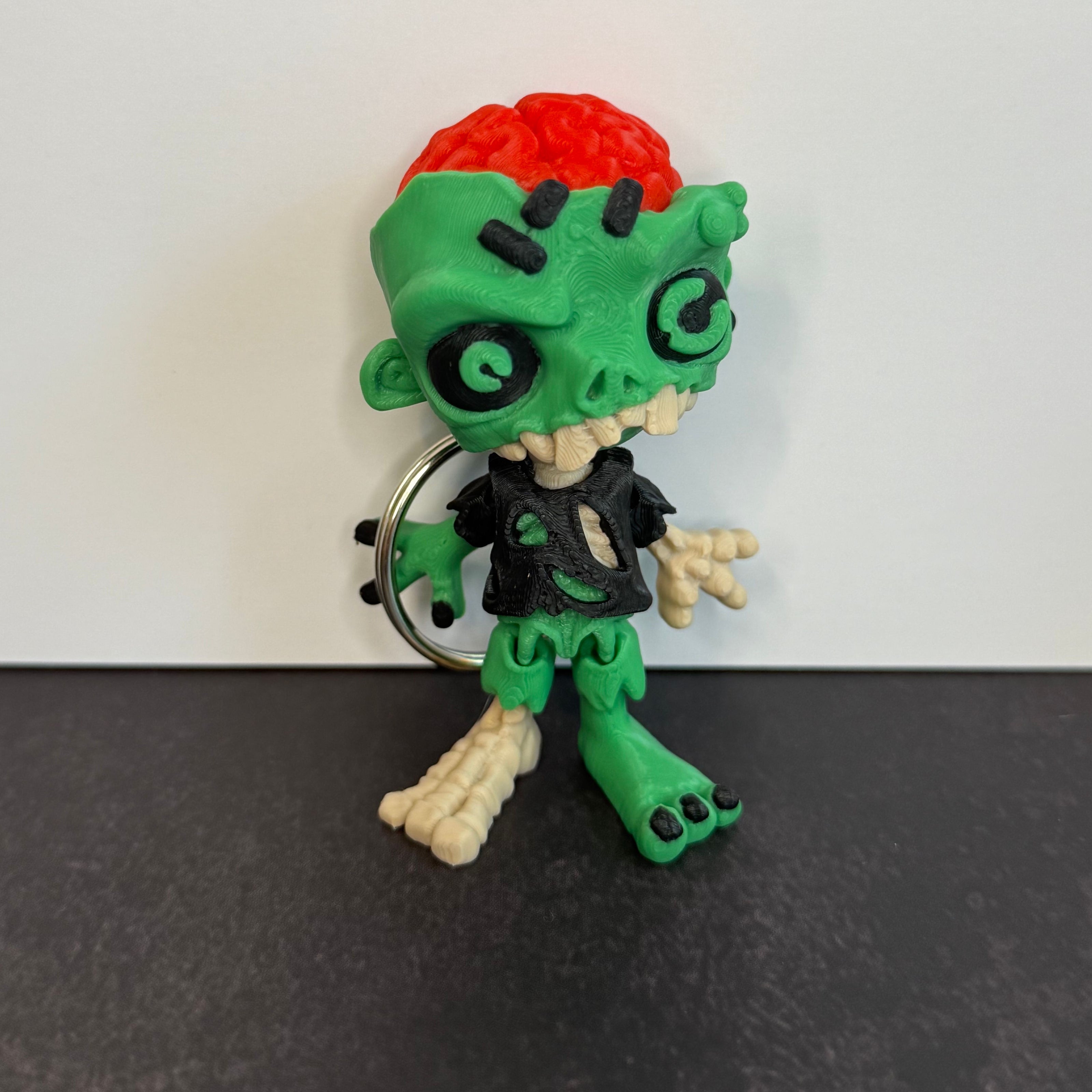 Zombie keychain with green skin, black eyes, and red brain on a black background