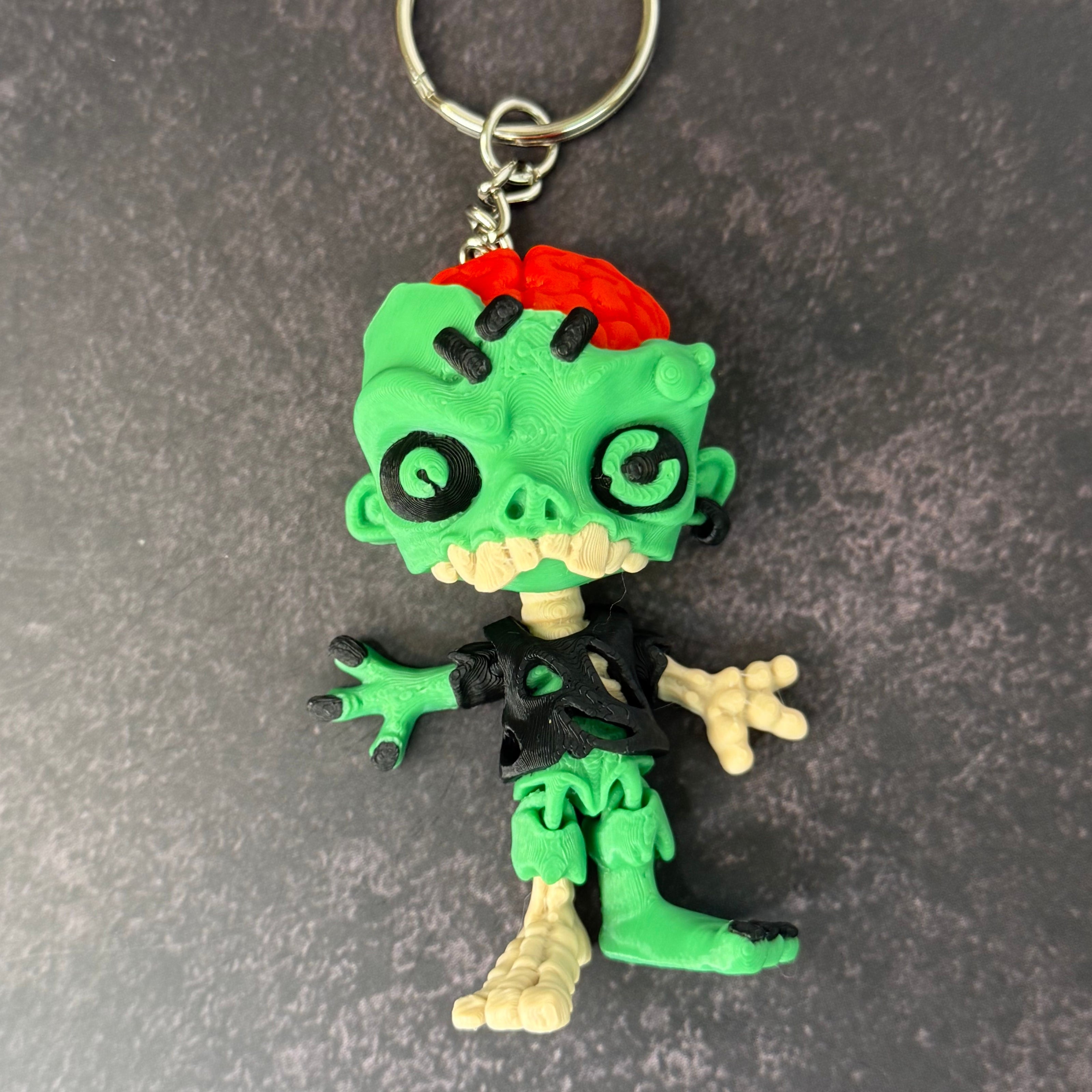 Zombie keychain with green skin, black eyes, and red brain on a black background
