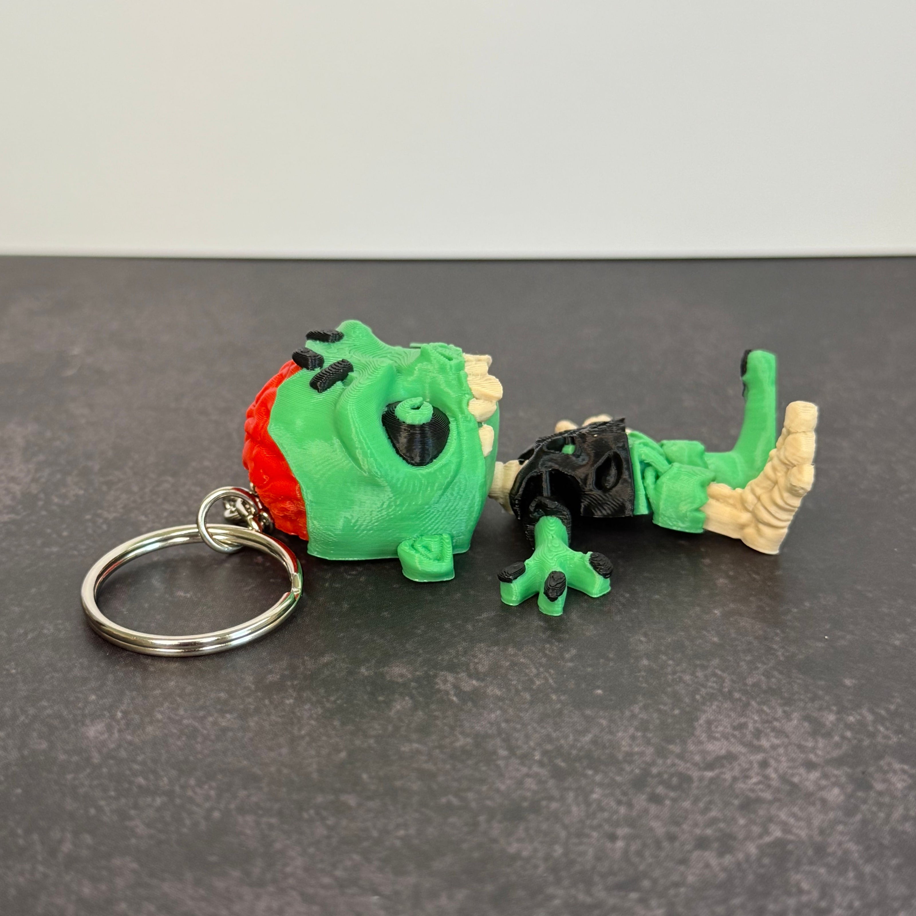 Zombie keychain with green skin, black eyes, and red brain on a black background