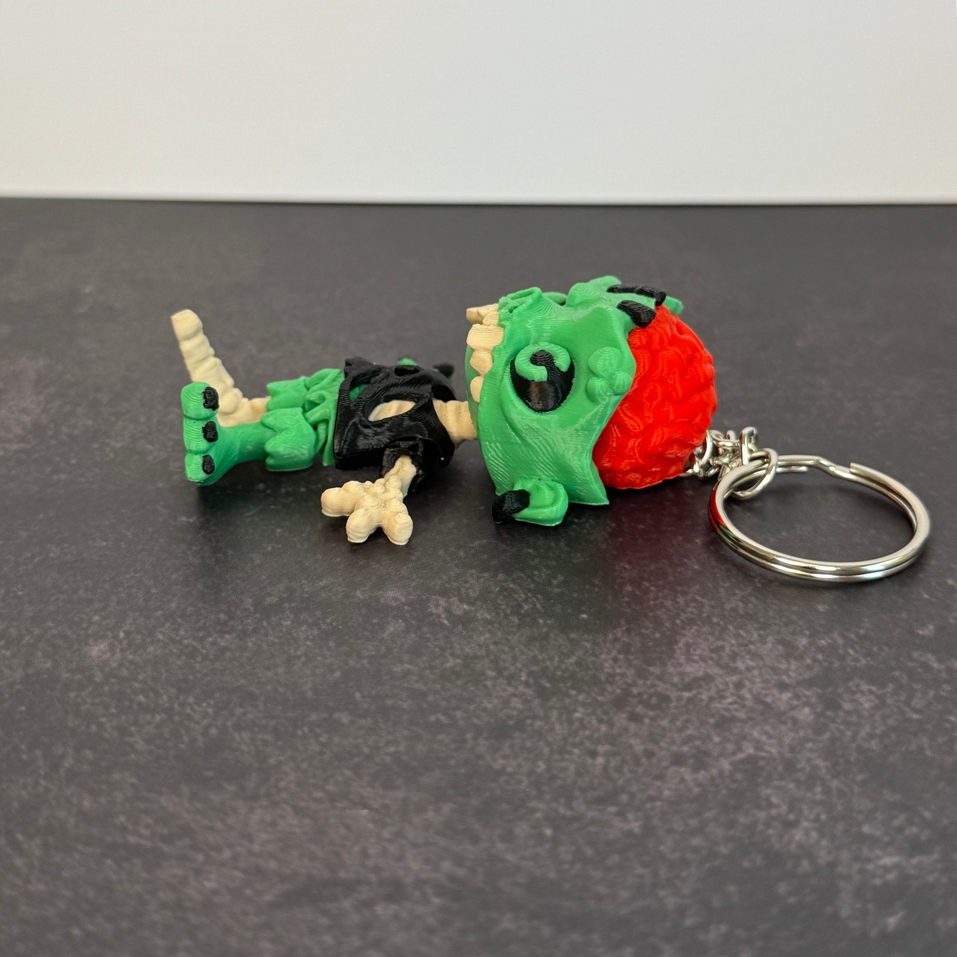 Zombie keychain with green skin, black eyes, and red brain on a black background