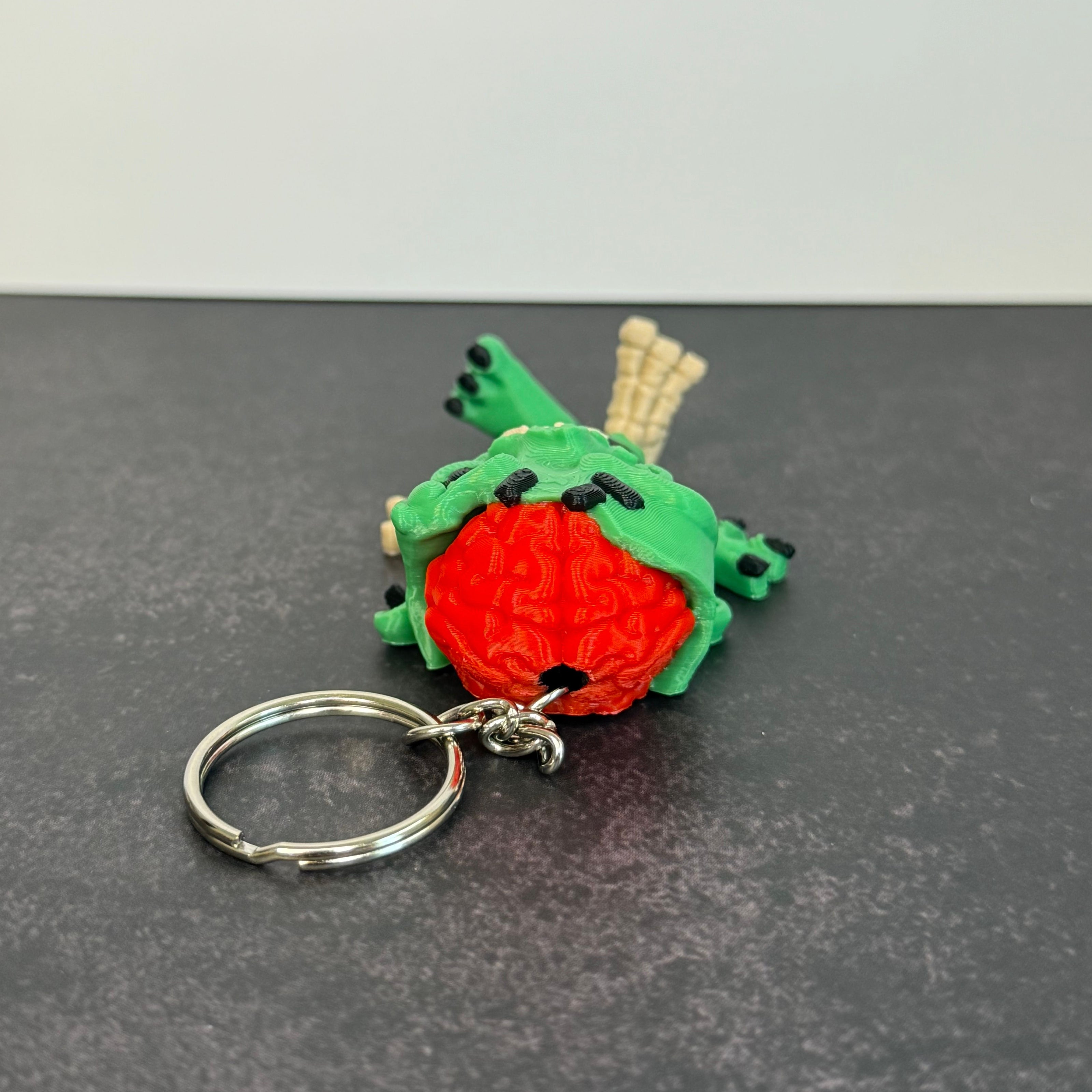 Zombie keychain with green skin, black eyes, and red brain on a black background