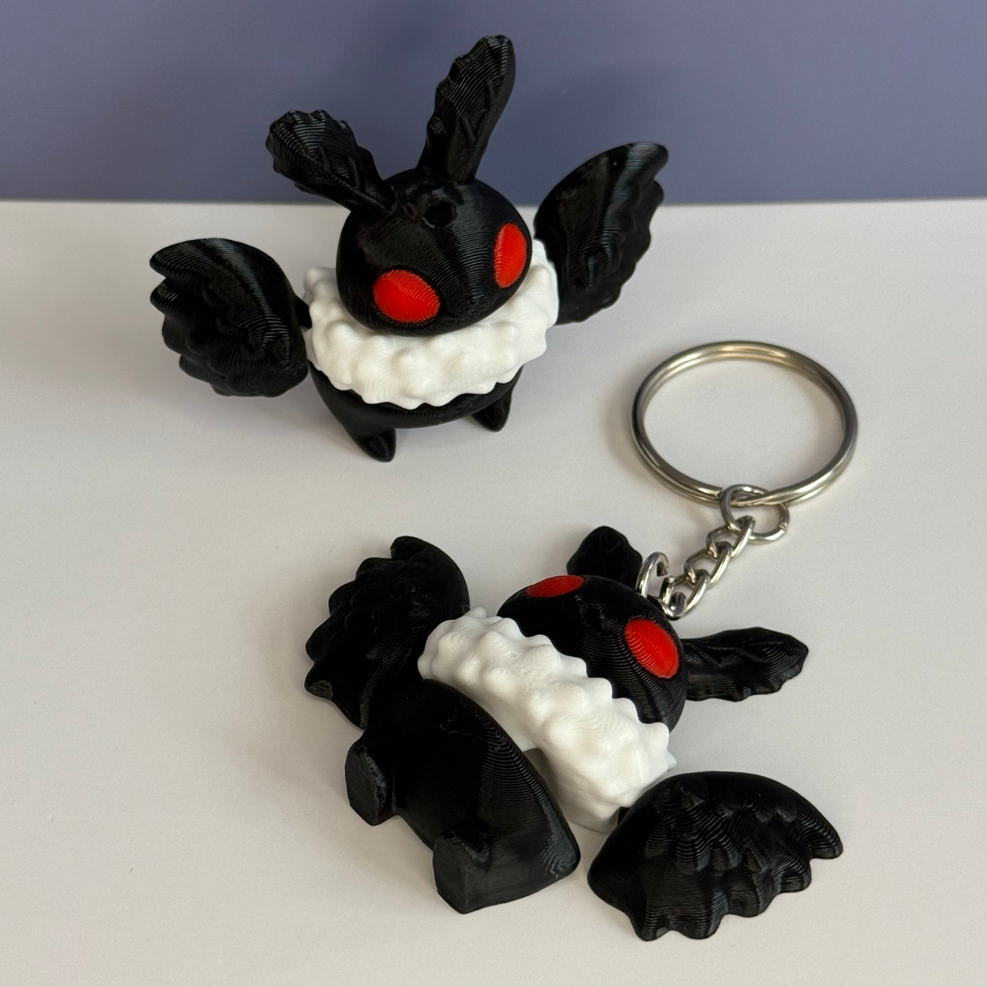 Black and white character figurine with red eyes and black wings on a white surface, one attached to a keychain.