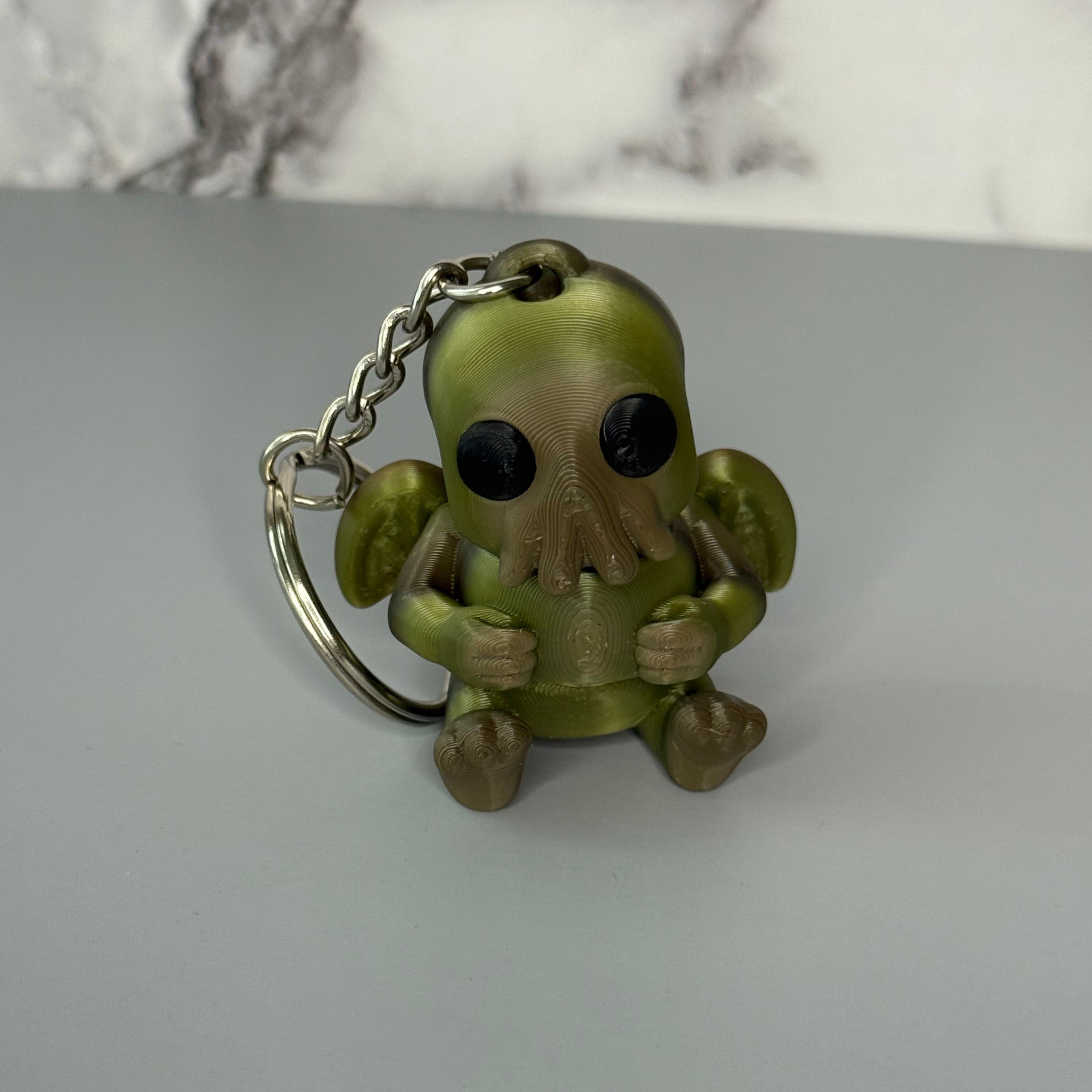 Sitting Cthulhu-shaped keychain figure with wings and face-tentacles on a gray background