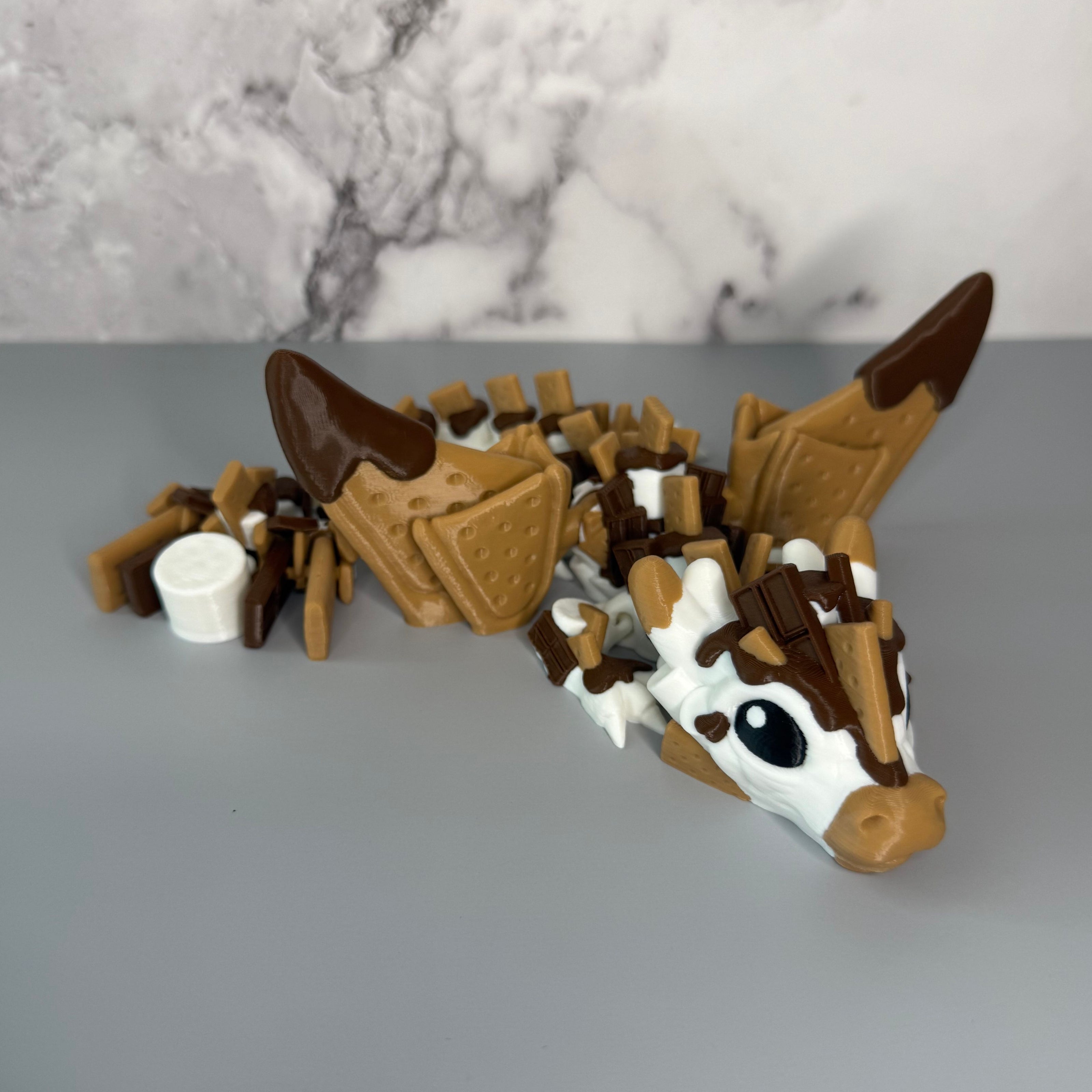Toy figure of a dragon with a white, brown, and tan body shaped liked marshmallows, chocolate, and graham crackers on a gray background