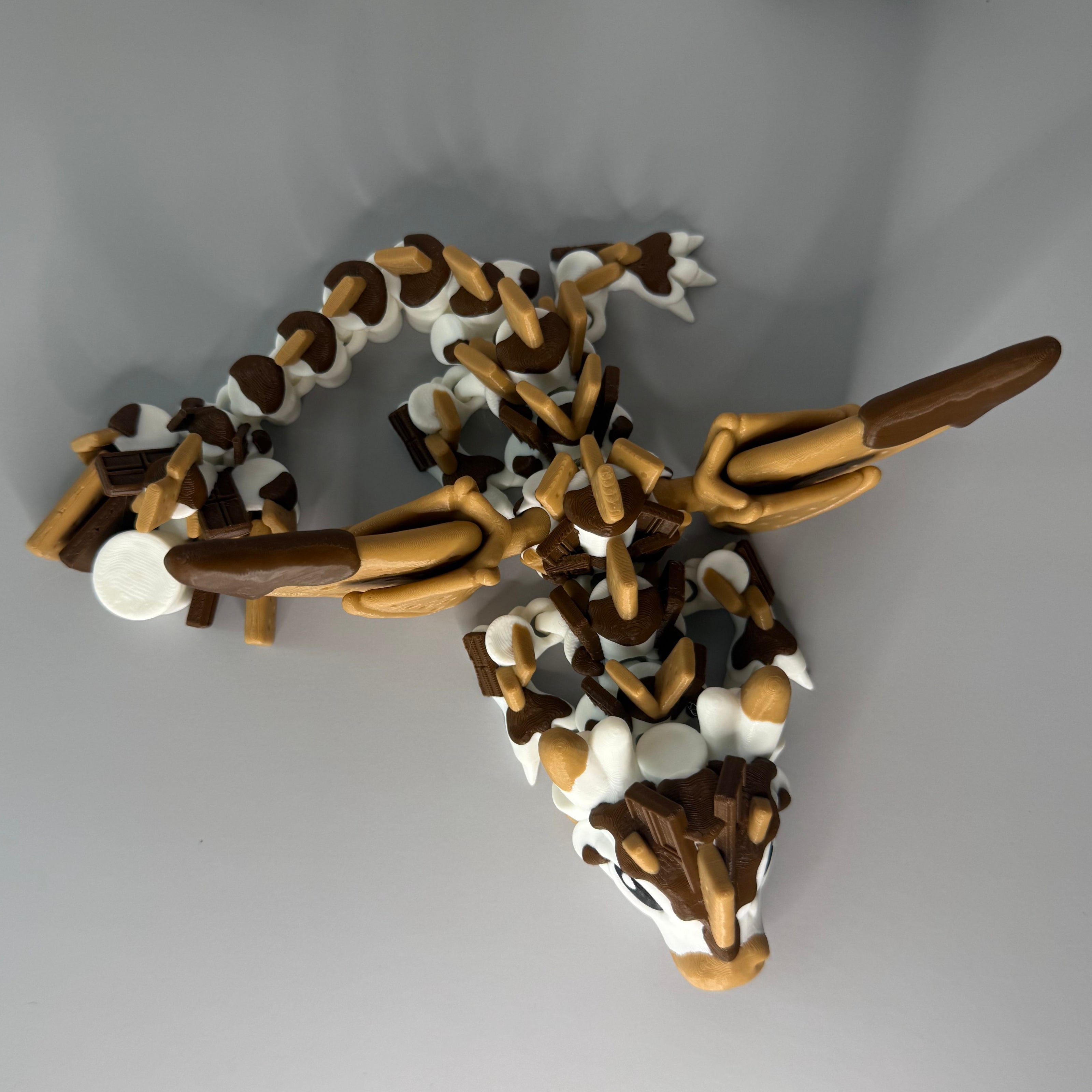 Top side of a toy figure of a dragon with a white, brown, and tan body shaped liked marshmallows, chocolate, and graham crackers on a gray background