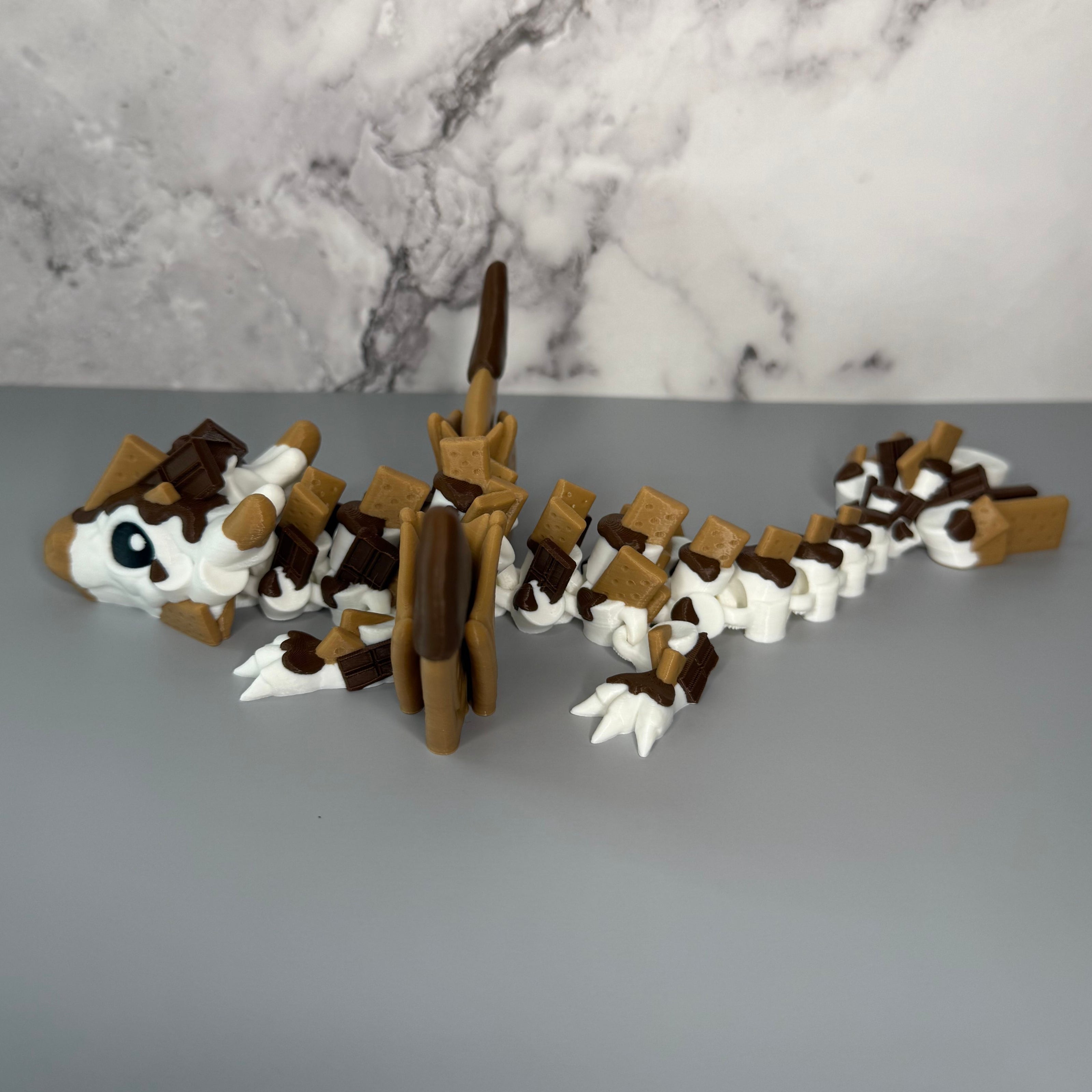 Side of a toy figure of a dragon with a white, brown, and tan body shaped liked marshmallows, chocolate, and graham crackers on a gray background