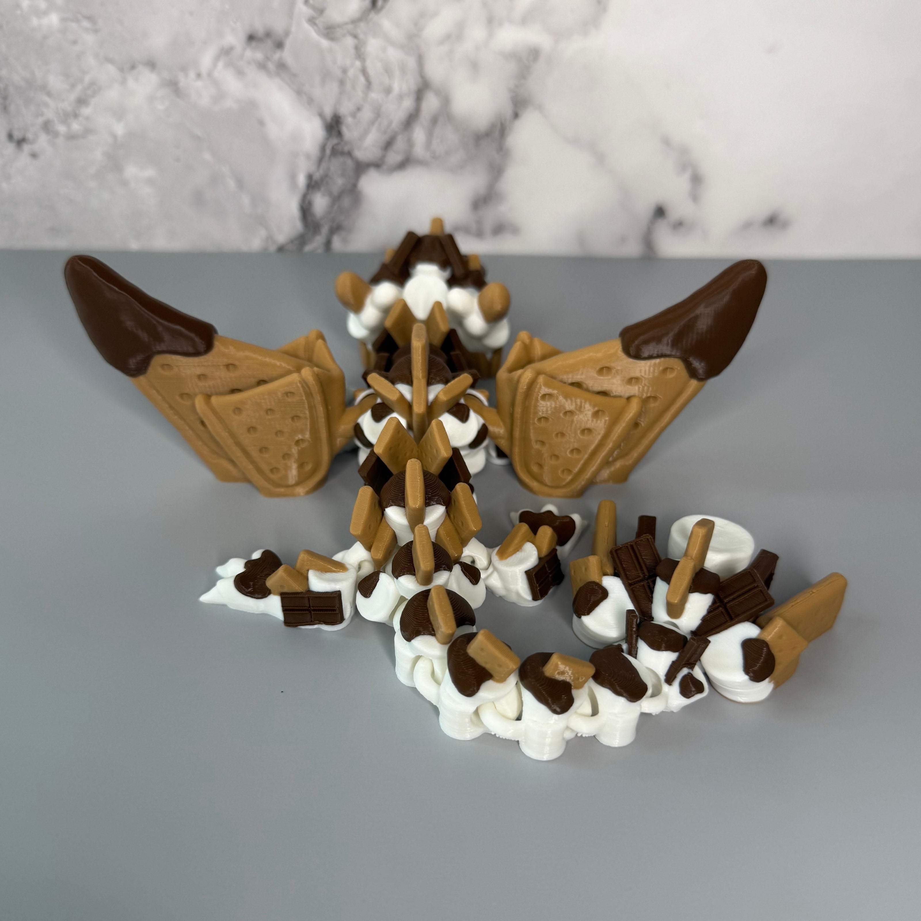 Back side of a toy figure of a dragon with a white, brown, and tan body shaped liked marshmallows, chocolate, and graham crackers on a gray background