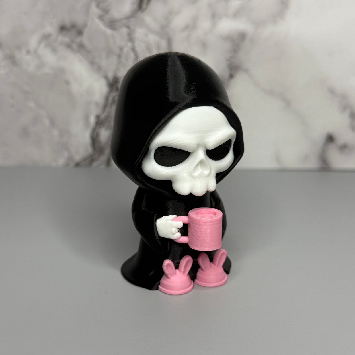 Skull figure in a black hood holding a pink mug with bunny feet slippers on a gray surface.
