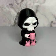 Skull figure in a black hood holding a pink mug with bunny feet slippers on a gray surface.