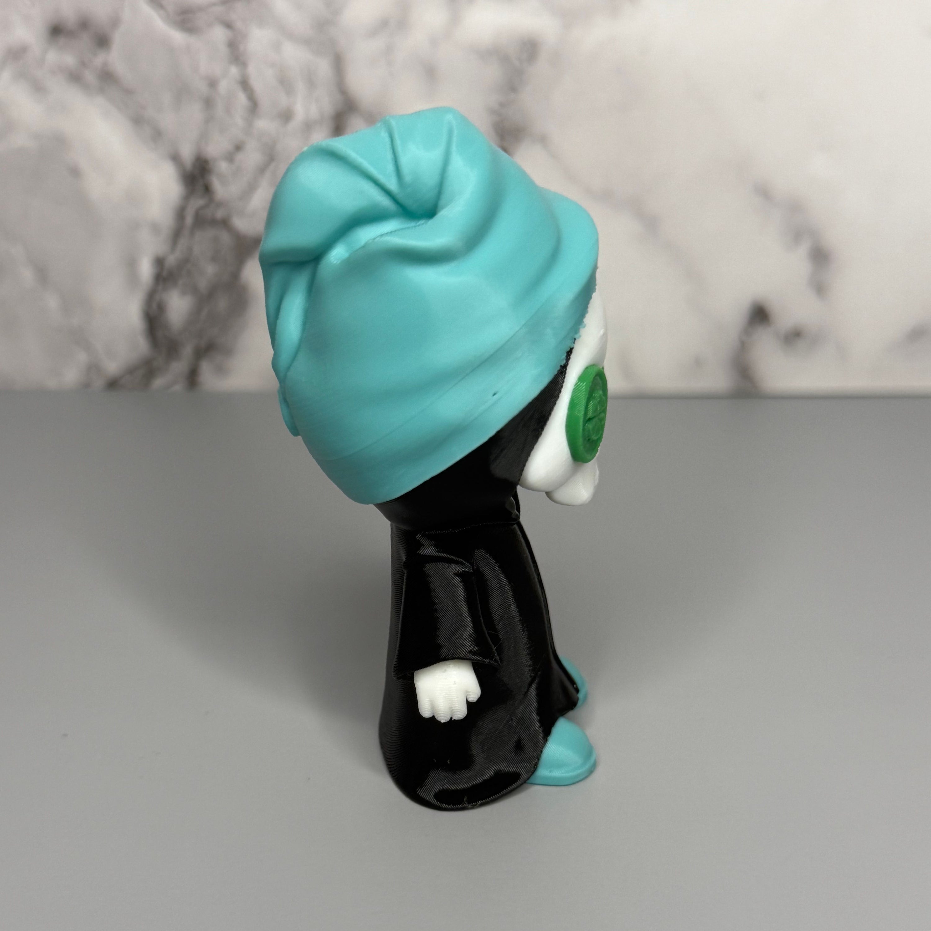 Left side of grim reaper figure with teal head towel and shoes, black cloak, and green discs on eyes on a gray marble background