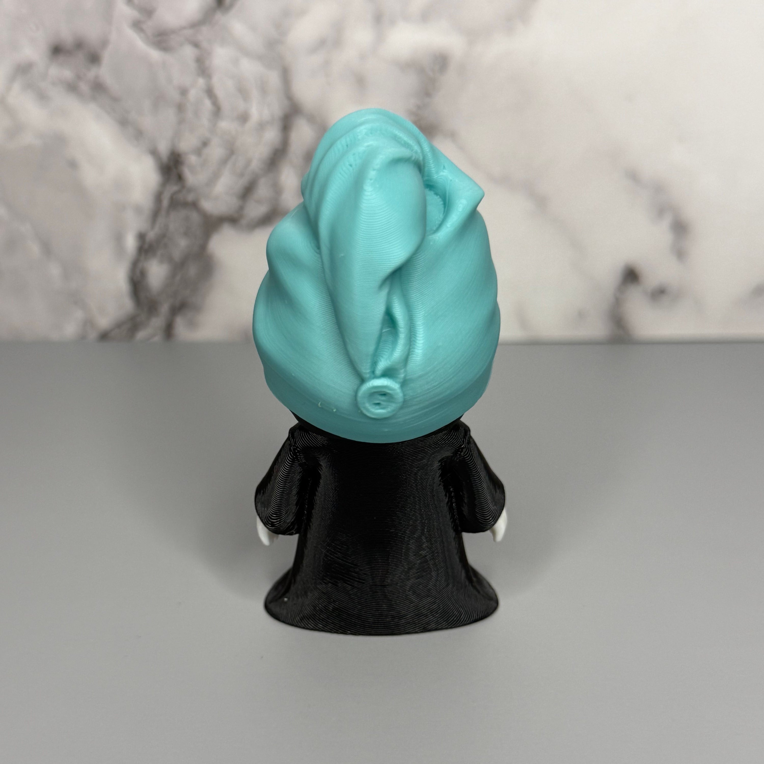 Back side of grim reaper figure with teal head towel and shoes, black cloak on a gray and marble background