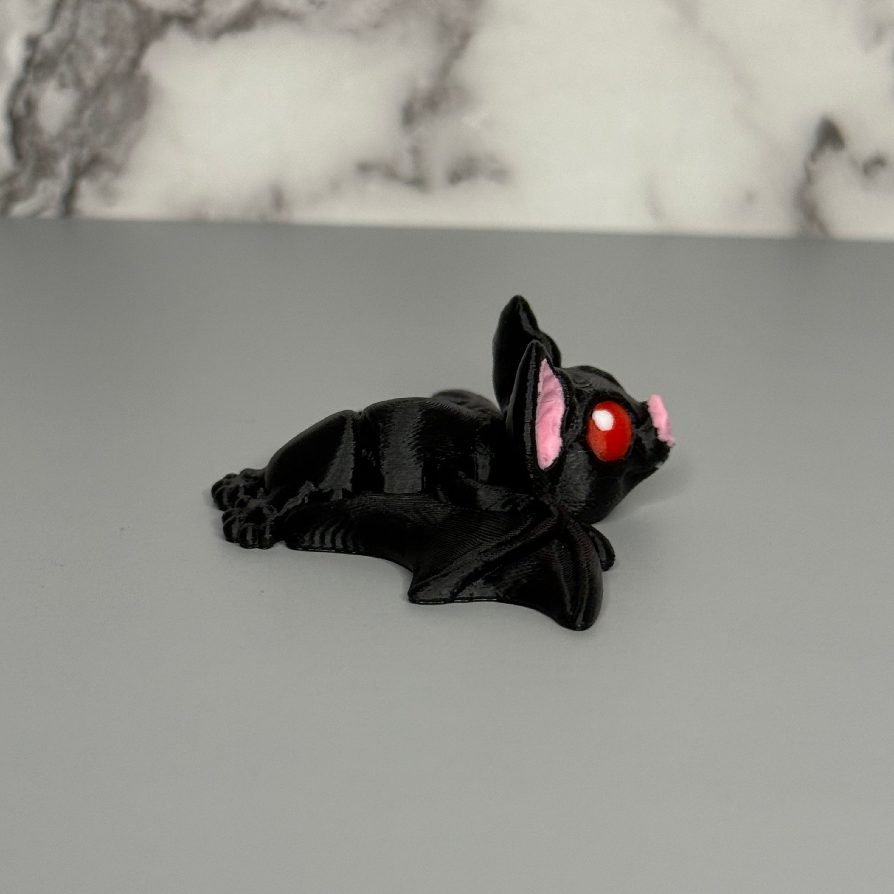 Left side of black bat figurine with red eyes and pink ears on a gray surface with marble background