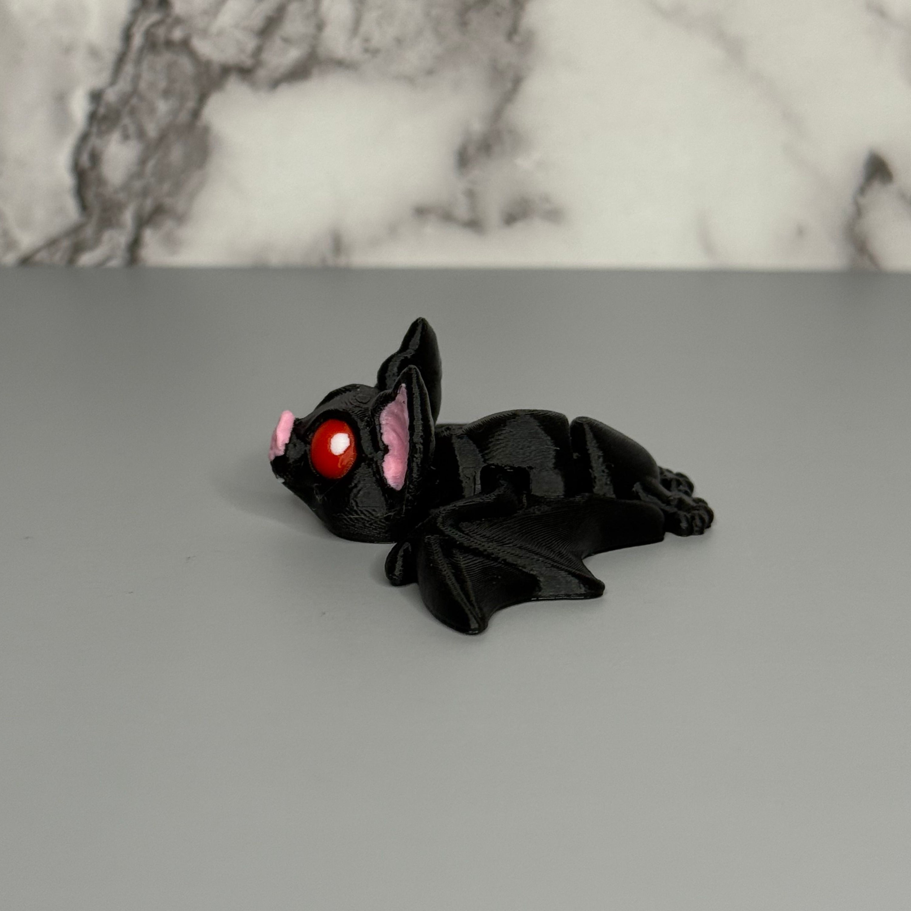 Right side of black bat figurine with red eyes and pink ears on a gray surface with marble background