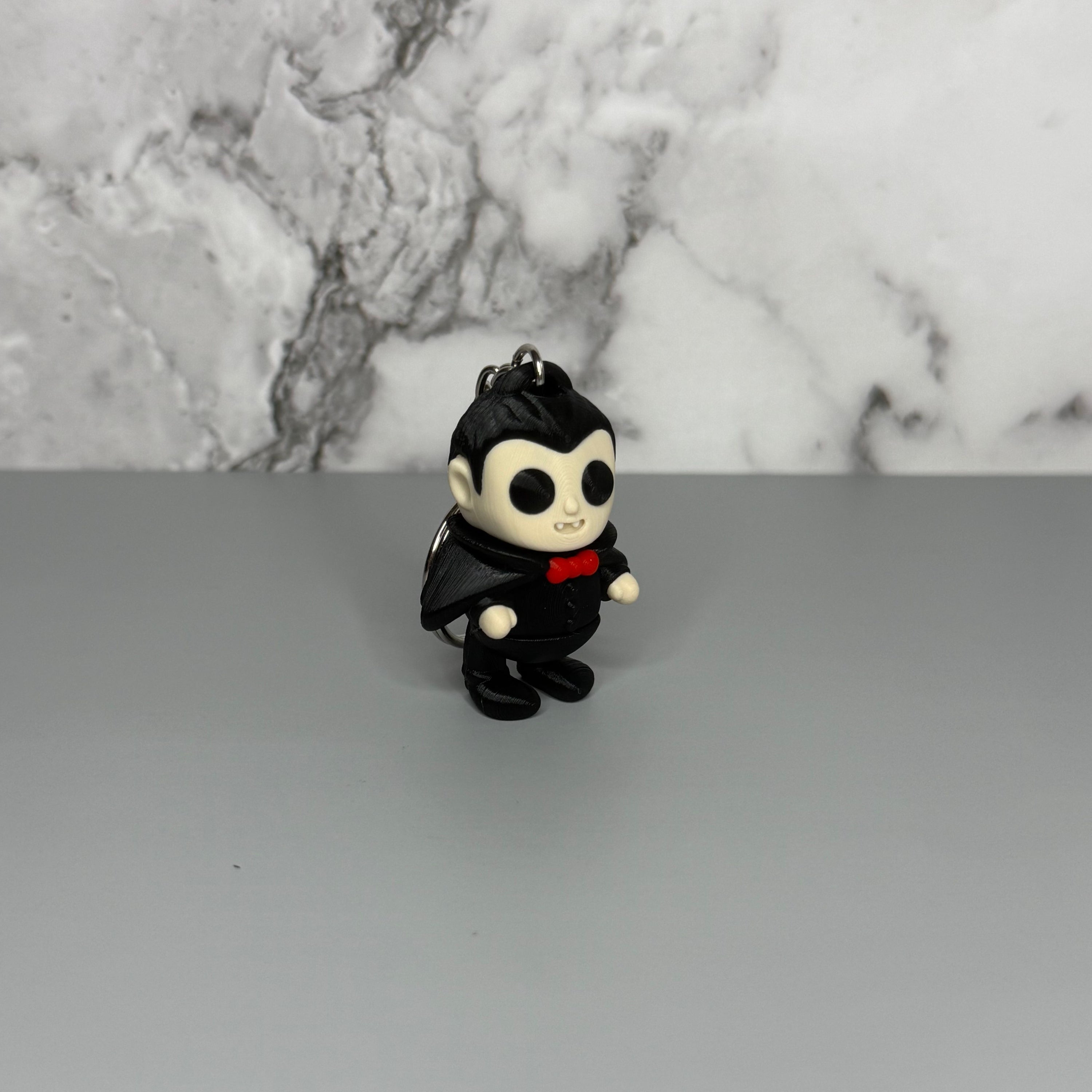 Keychain of a dracula vampire figure with a black cape, white fangs, and a red bowtie on a gray marble background